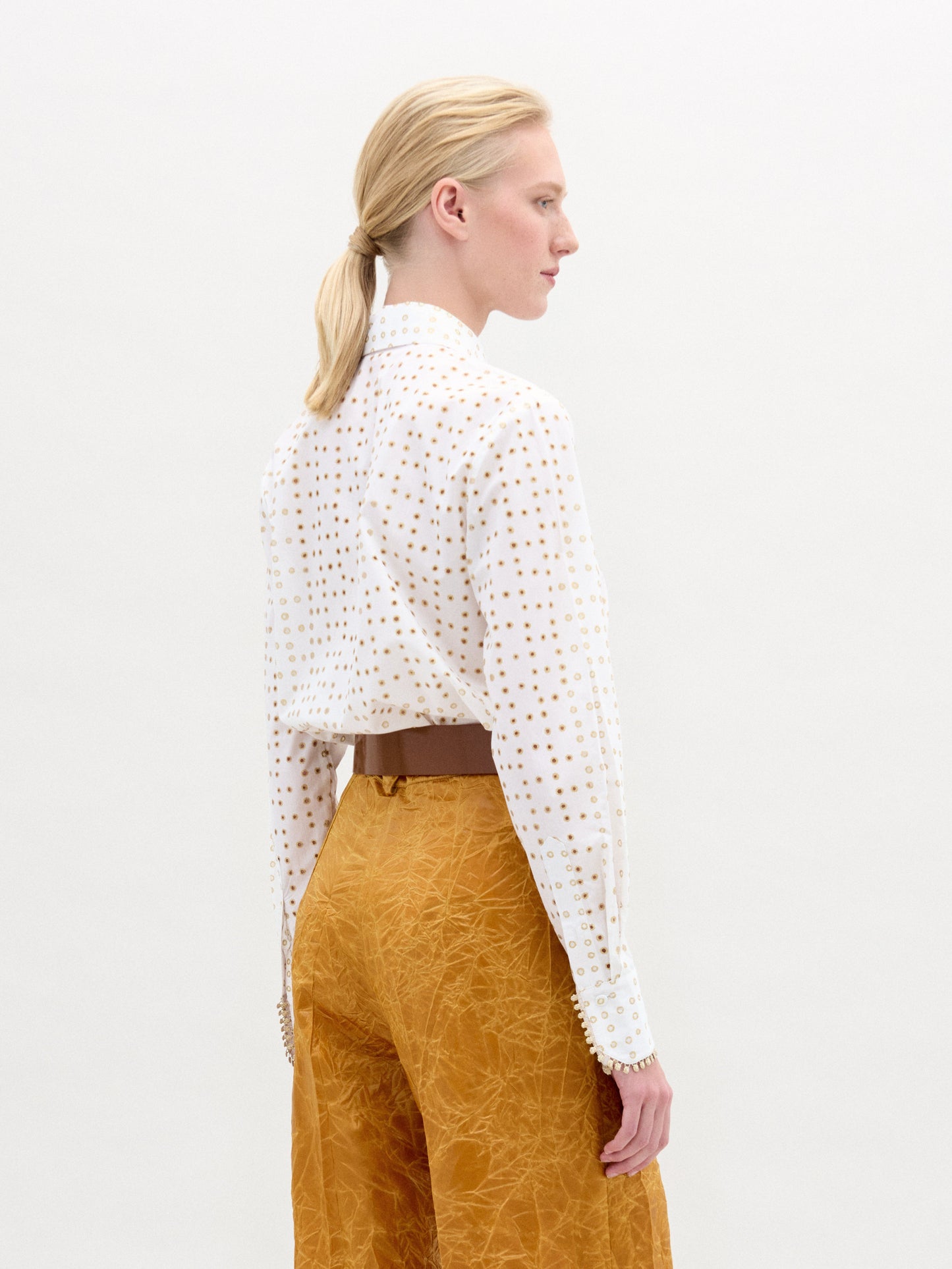 Una persona con el pelo rubio recogido en una coleta baja, de pie y de lado, lleva la blusa Naroa -una blusa de manga larga de tela italiana con lunares dorados- combinada con unos pantalones mostaza texturizados. Fabricada en Colombia, se muestra sobre un sencillo fondo blanco.