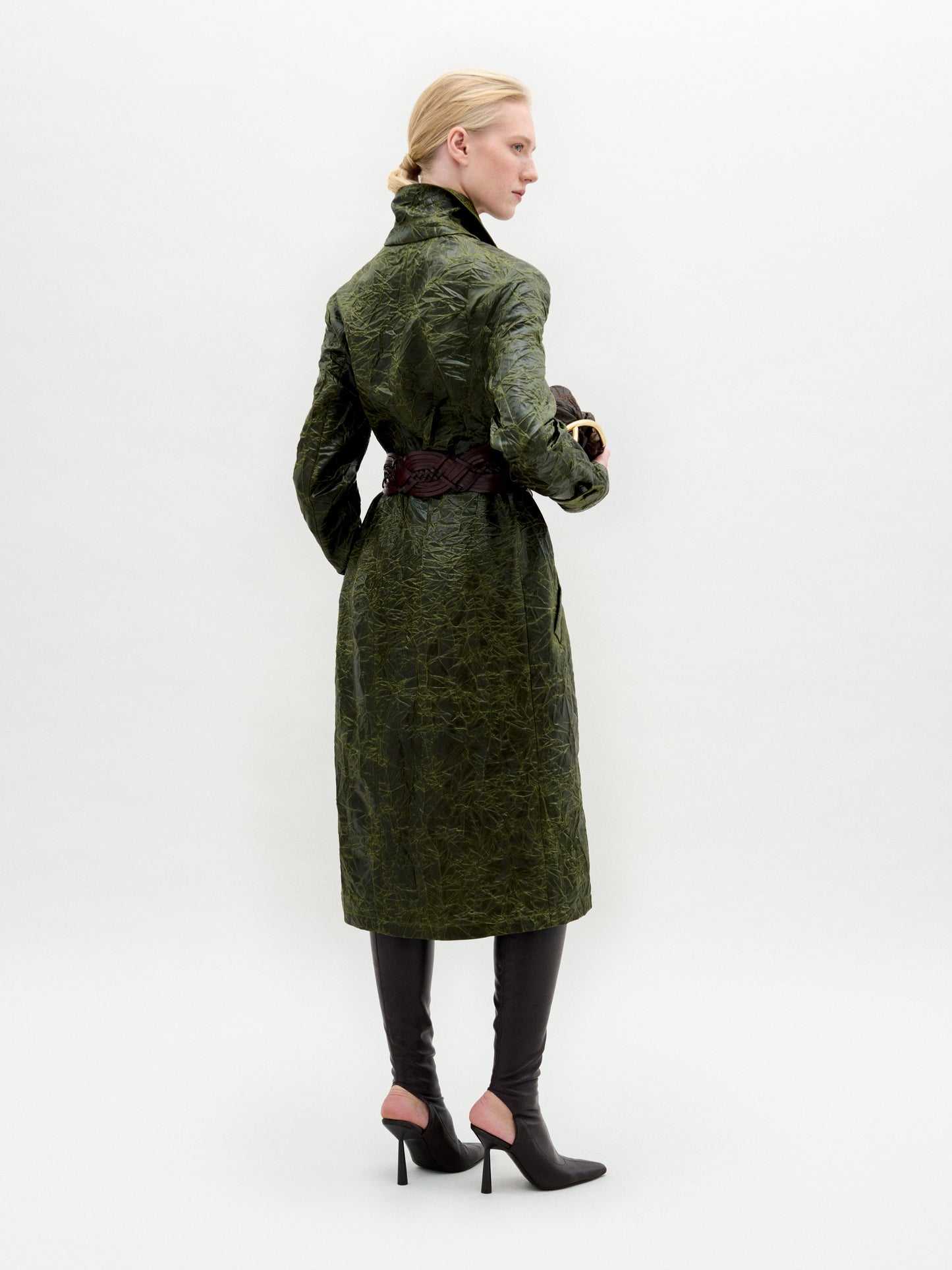 Una persona, de espaldas, lleva el Nilo Trench -un abrigo verde texturizado fabricado en Colombia- ceñido a la cintura, combinado con botas negras de tacón y guantes marrón oscuro. El fondo es liso y de color claro.