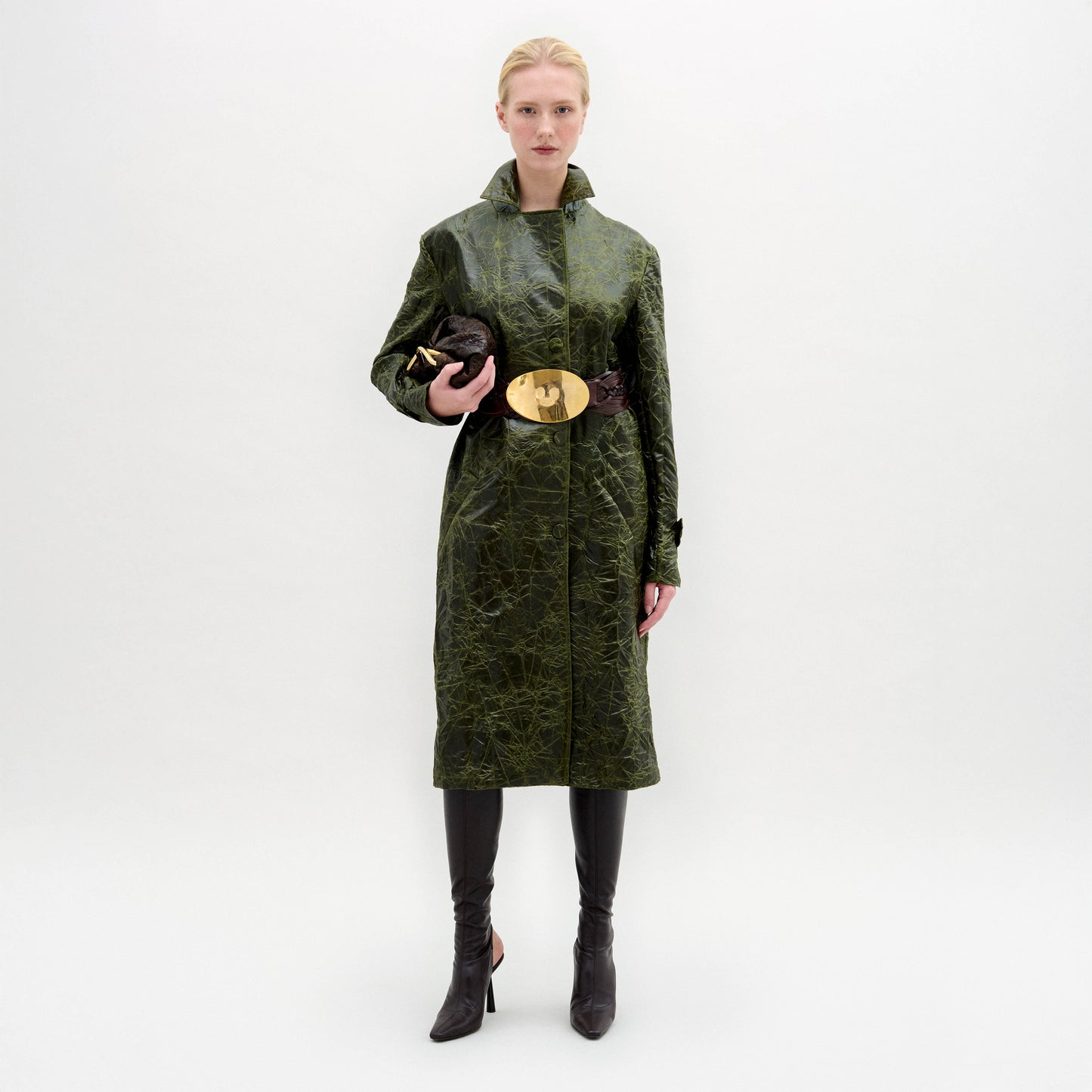 Una persona sobre un fondo blanco liso lleva el modelo Nilo Trench verde oscuro texturizado con un cinturón ancho dorado, pantalones oscuros, botas puntiagudas y un bolso de mano oscuro. El look lleva tela italiana y está hecho en Colombia.