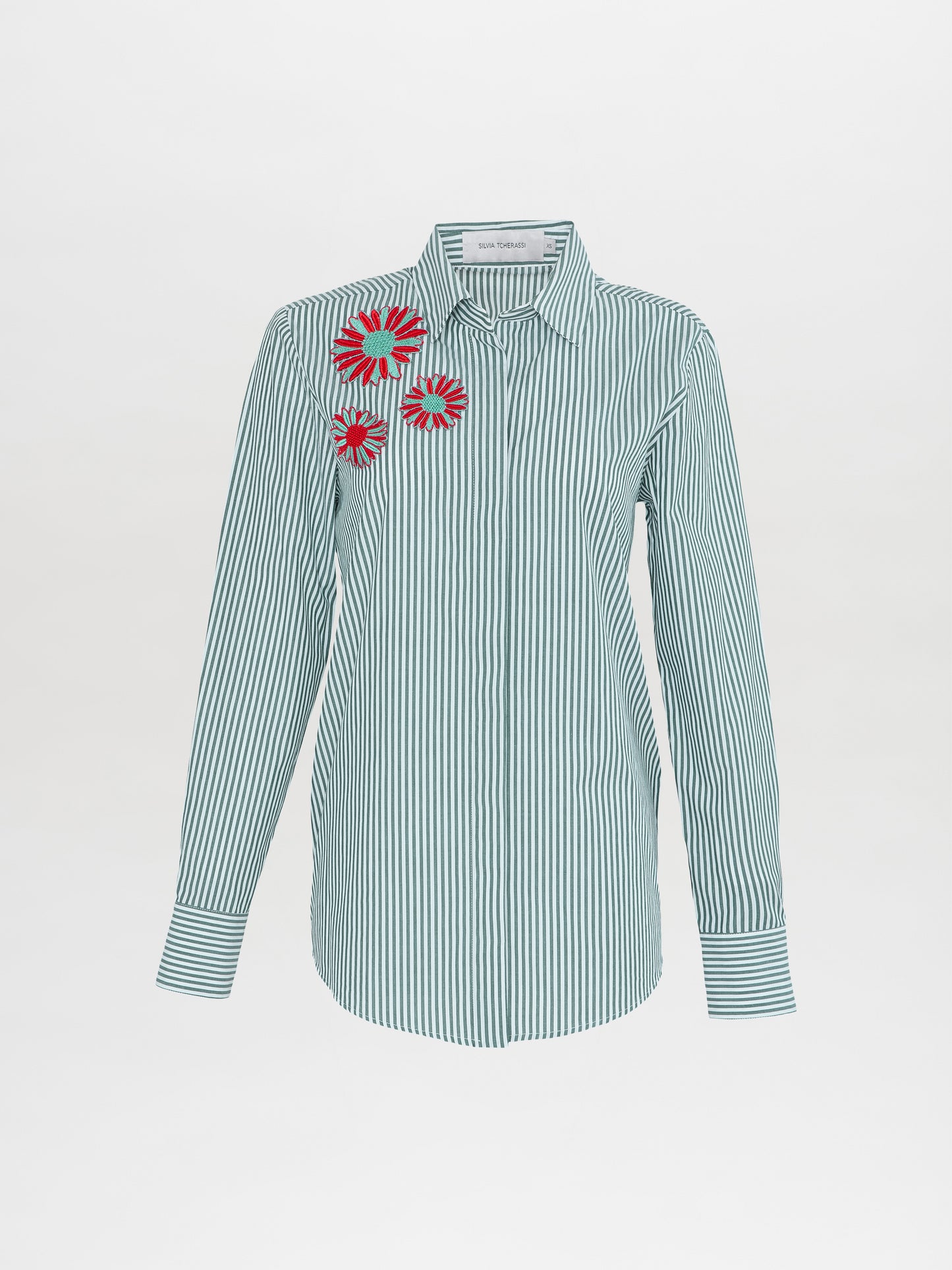 La blusa Naroa de rayas verdes es una camisa de manga larga con botones, rayas verticales verdes y blancas y tres parches circulares rojos y blancos en el hombro izquierdo.