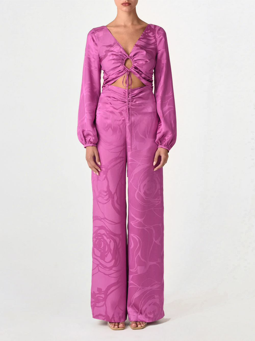 Una persona mira al frente y lleva el Blusa Odette Jacquard Orquídea Magenta, un top magenta de manga larga con la parte central recortada y detalle de nudo delantero en sutil jacquard floral. Lo combinan con pantalones de pierna ancha y sandalias abiertas.