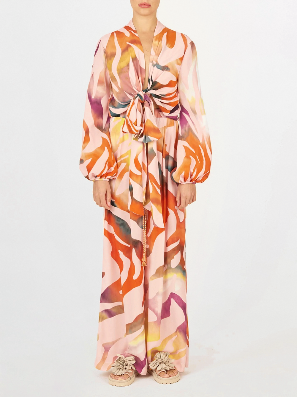 Una persona está de frente y lleva la Blusa Honey Blush Prismatic de Silvia Tcherassi, una blusa de manga larga con un colorido estampado abstracto en tonos rosa, naranja y morado. La acompañan unos zapatos planos con volantes sobre un fondo blanco liso.