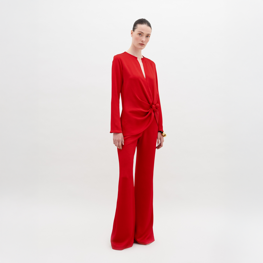 Una persona está parada sobre un fondo blanco y viste un atuendo rojo, que incluye una camiseta de manga larga con detalle de lazo en la cintura y el Pantalón Palermo. El estilo tiro alto complementa su cabello oscuro, que está recogido hacia atrás, mientras mira a la cámara de manera neutral.