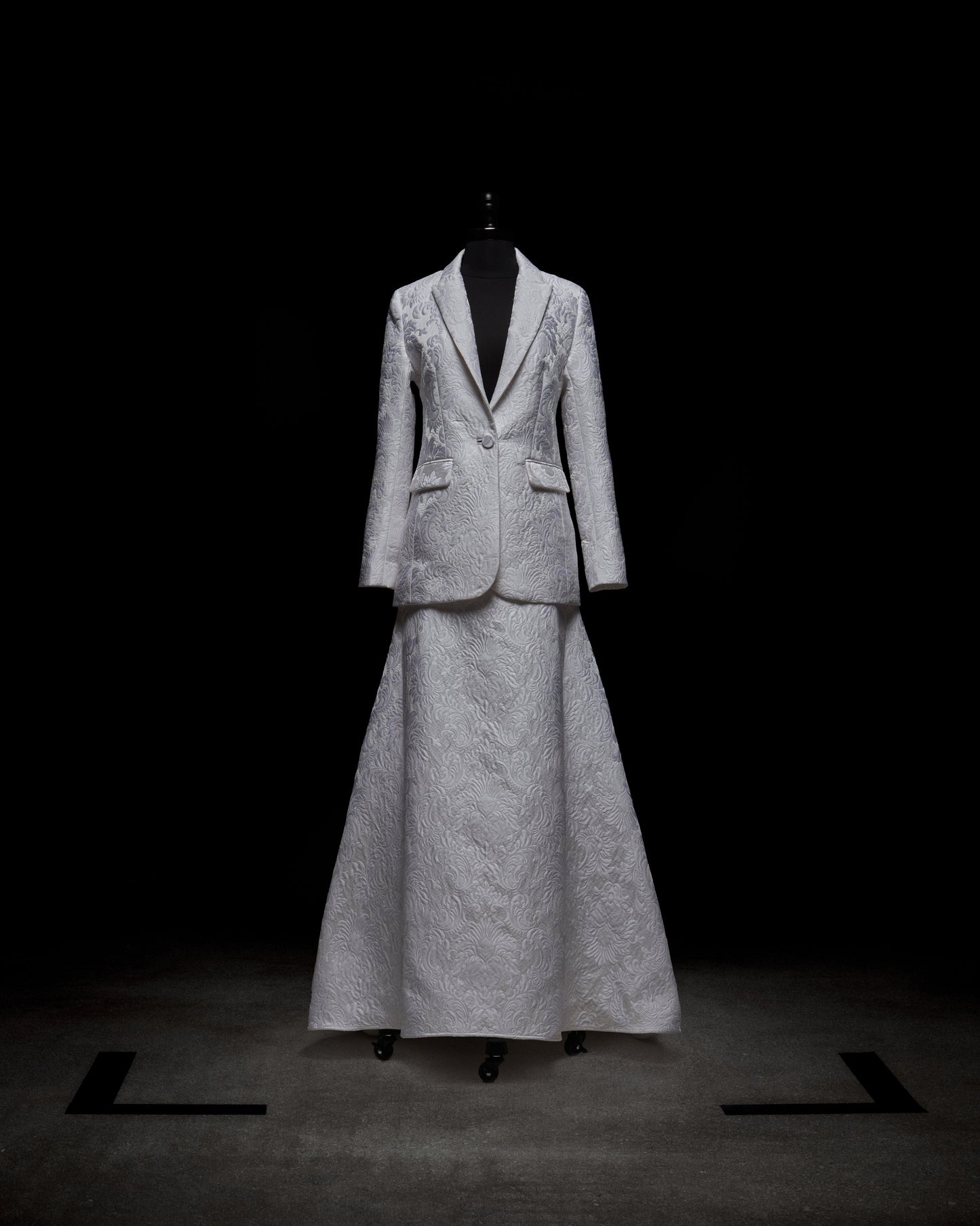 La falda Imola, un vestido de traje blanco texturizado, se exhibe sobre un maniquí negro sobre un fondo oscuro. Incluye un blazer ajustado con solapas de muesca y una falda larga y vaporosa.