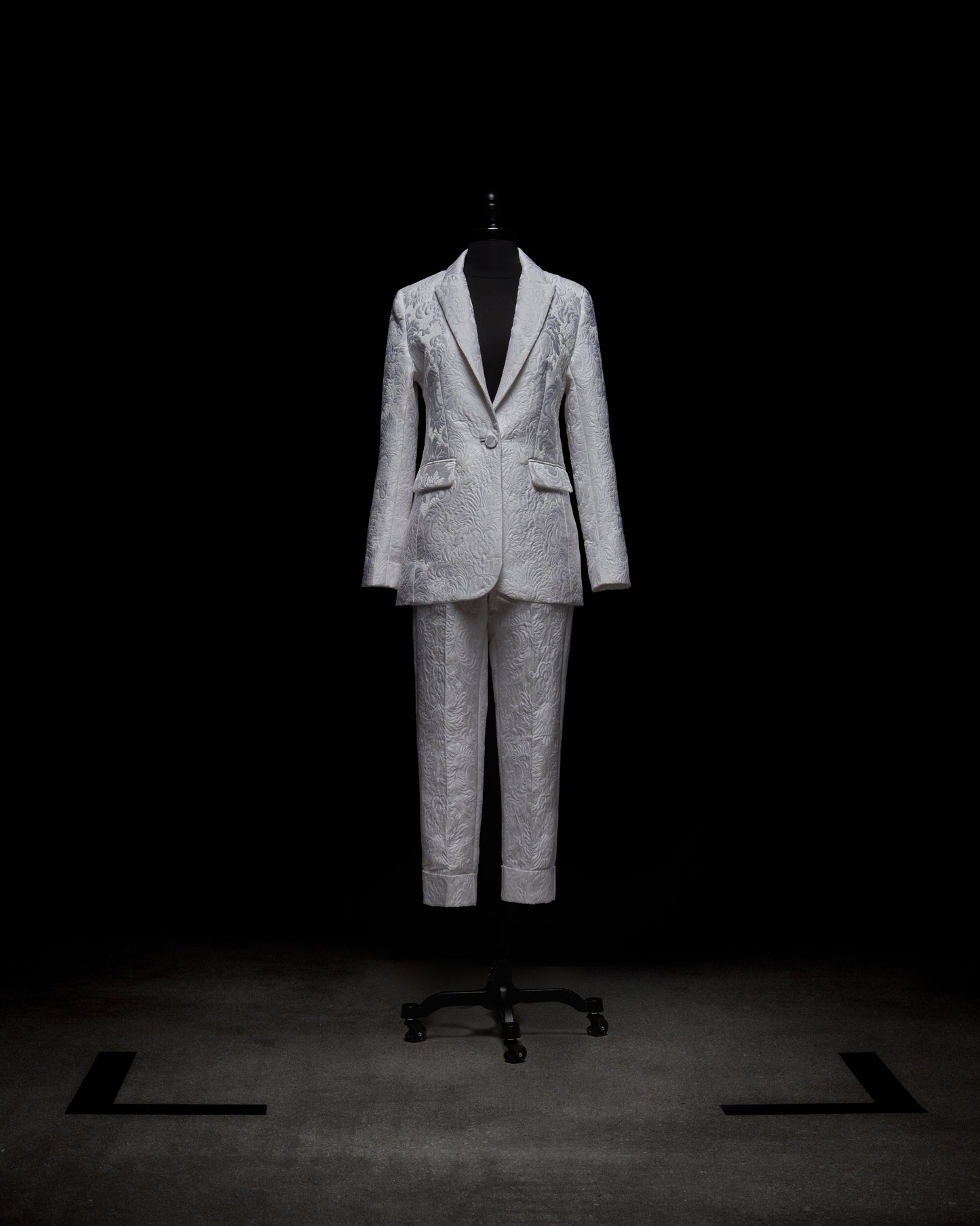 Sobre un maniquí negro y fondo oscuro, se exhibe el pantalón Moad, parte de un traje gris estampado con chaqueta con botones y bolsillos, y pantalones a juego. La atención se centra en la textura y los detalles del diseño.