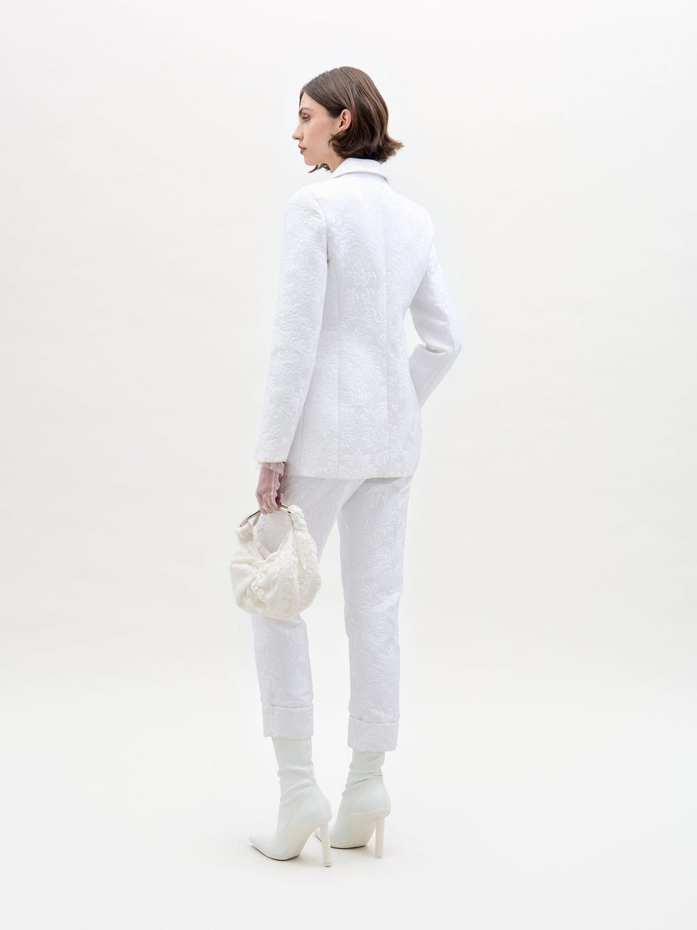 Una persona con cabello corto mira hacia otro lado, vistiendo un traje de pantalón Moad blanco con textura, puños remangados y botas blancas de tacón alto, y sosteniendo una bolsa blanca en su mano derecha contra un fondo blanco liso.