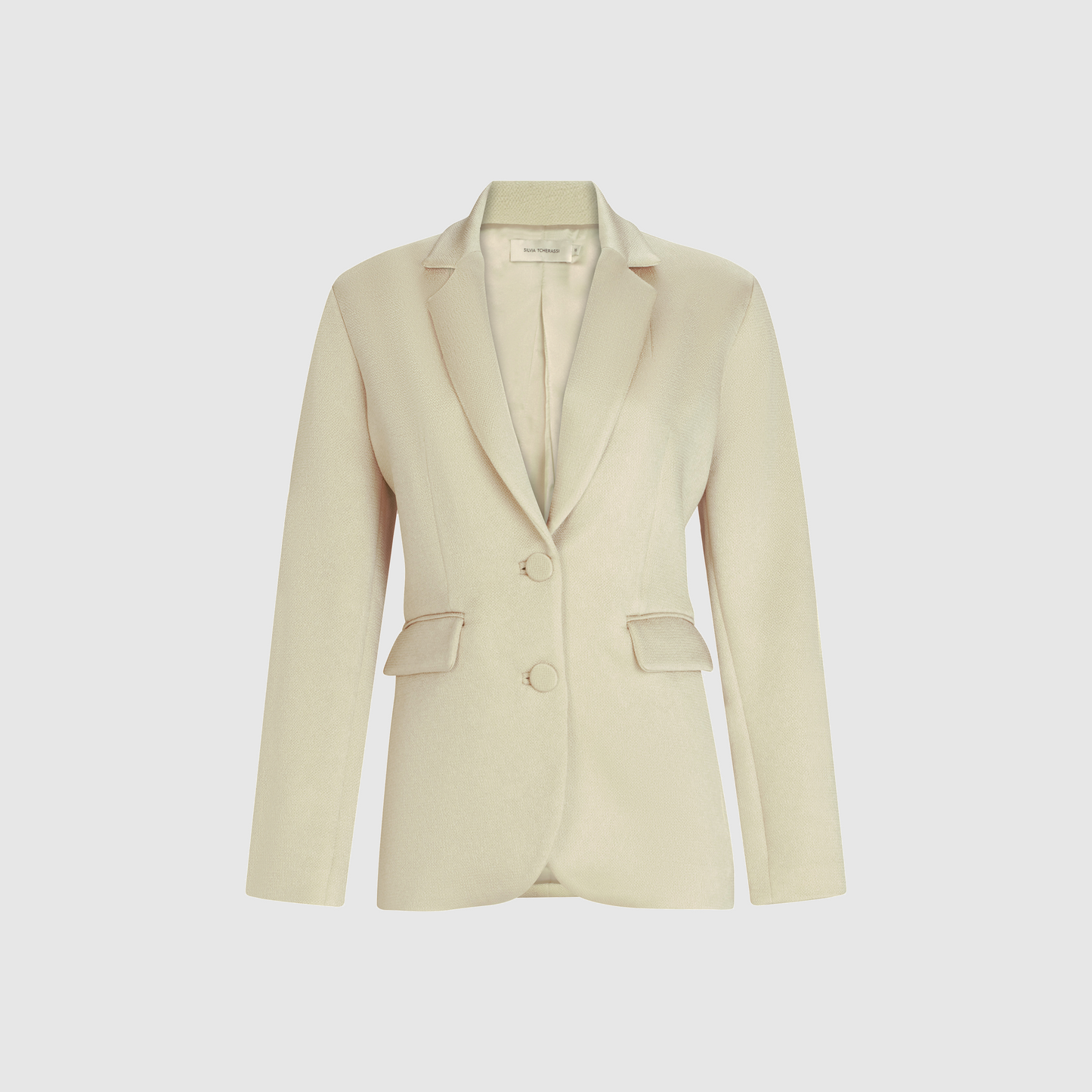 Chaqueta Rebeca para mujer, de color beige claro, de corte entallado, con mangas largas, solapas con muescas, dos bolsillos con solapa delanteros y dos botones delanteros en satén martillado, que se muestra sobre un fondo blanco liso.