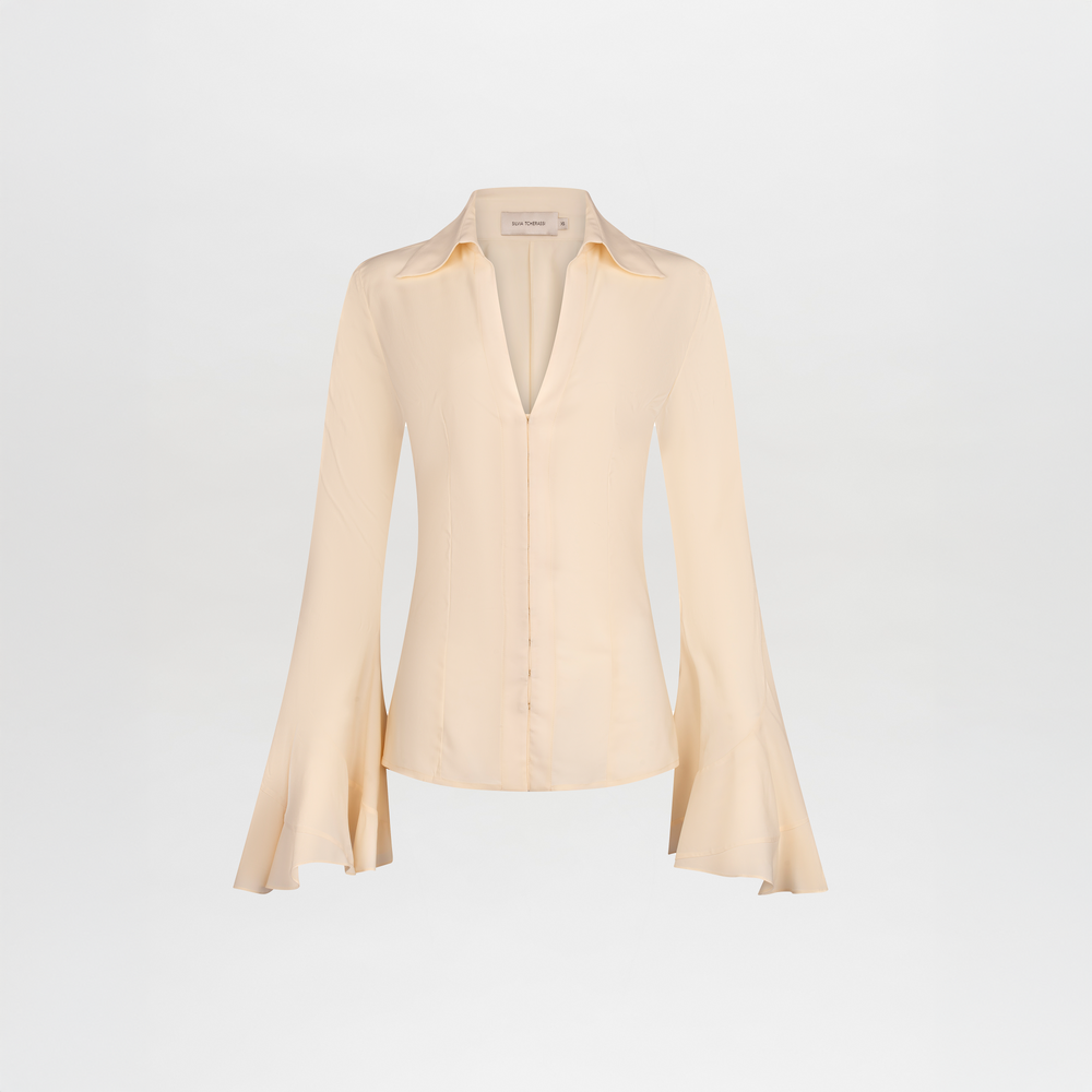 La Blusa Reggie es una blusa beige claro de manga larga, confeccionada en suave seda, con silueta ajustada, escote en V pronunciado y puños acampanados. Fabricada en Portugal y presentada sobre un fondo blanco liso.