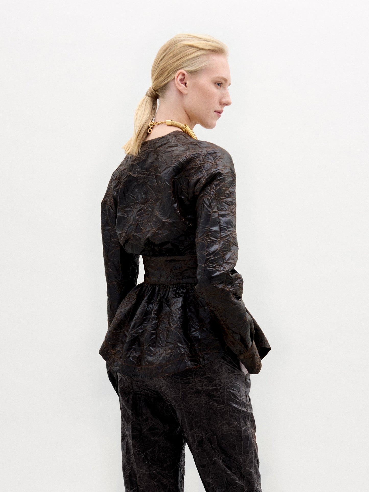 Una persona de piel clara y pelo rubio recogido en una coleta baja mira ligeramente hacia otro lado, modelando la blusa Saanvi en marrón oscuro con cinturón en la cintura y detalle de peplum, confeccionada con tejido italiano en Colombia, sobre un fondo blanco liso.