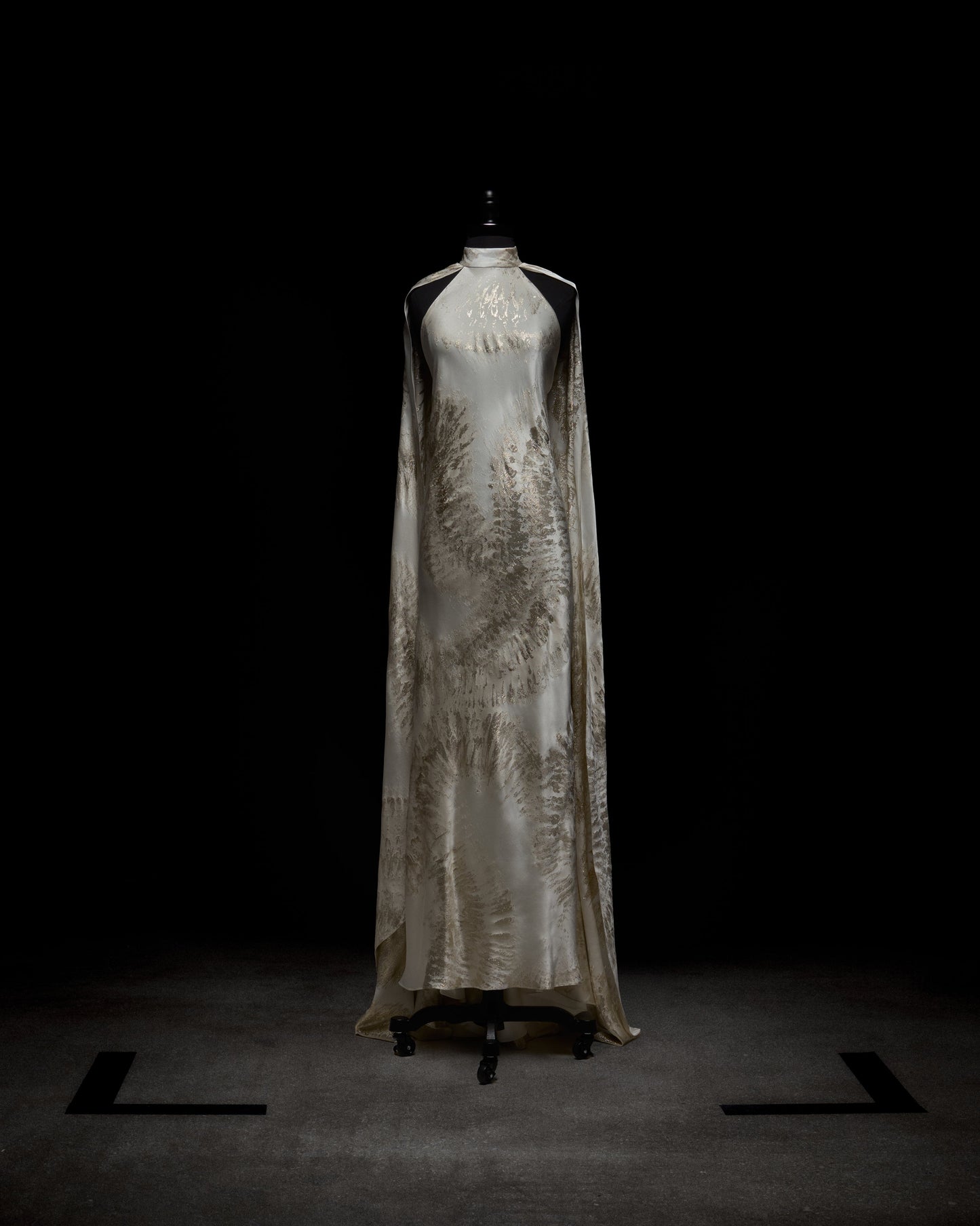 Vestido Sabina: Un vestido color marfil, largo hasta el suelo, sin mangas, con escote alto y sutiles patrones de hojas, exhibido en un maniquí negro sobre un fondo oscuro.