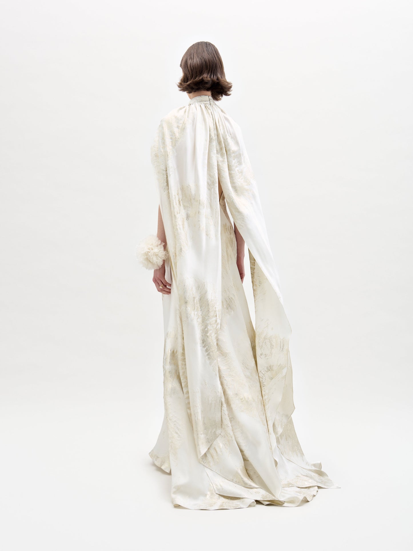 Una persona con un corte de pelo corto está de espaldas, luciendo el Vestido Sabina: un vestido de satén blanco hasta el suelo con capa y sutiles estampados texturizados. El fondo es blanco liso.