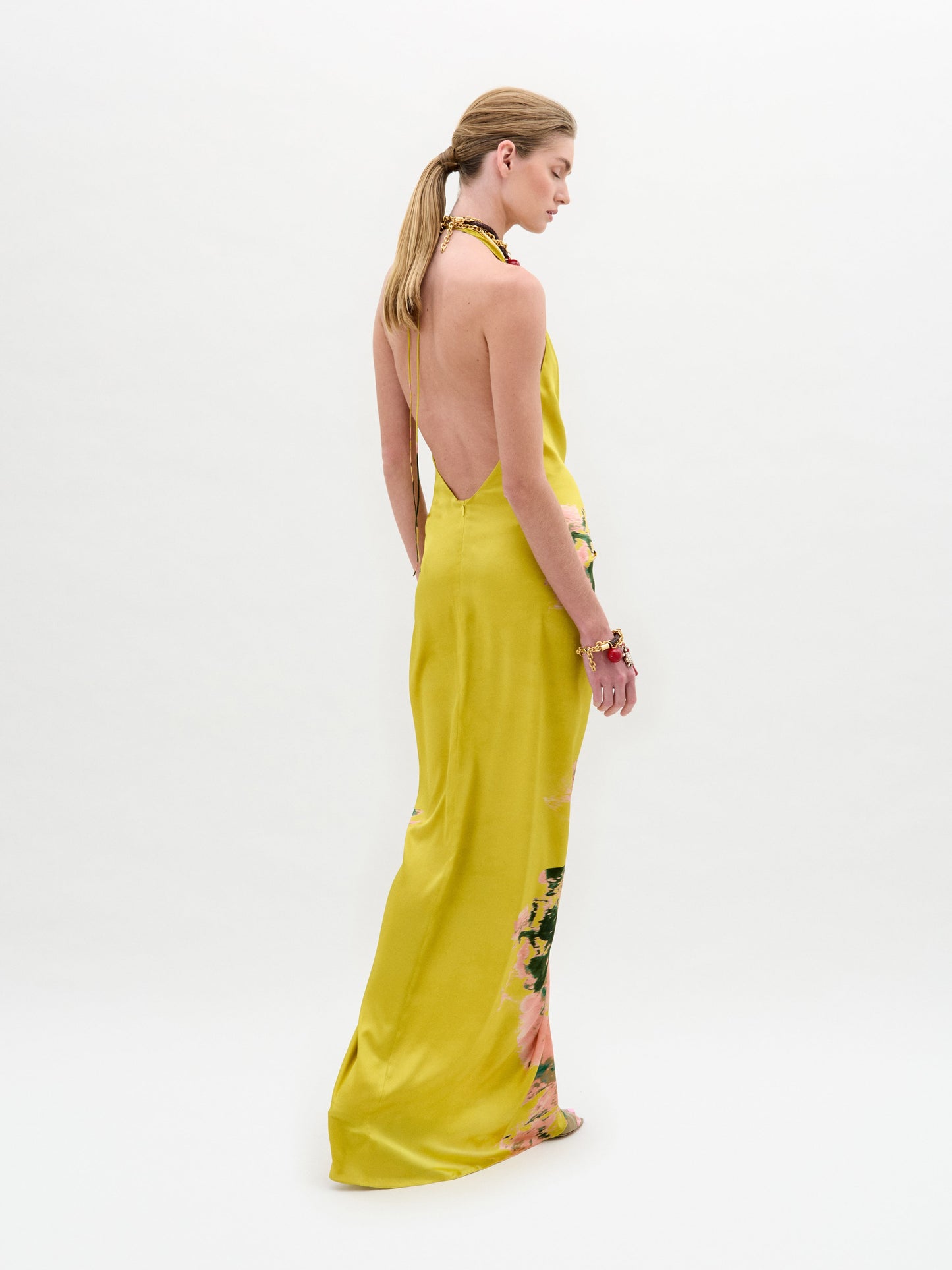Una persona viste de lado el vestido Jazmin, un midi sin mangas ni espalda con detalles florales en el bajo y un elegante escote halter. Lleva el pelo recogido en una coleta baja y brazaletes en una muñeca, sobre un fondo blanco liso.