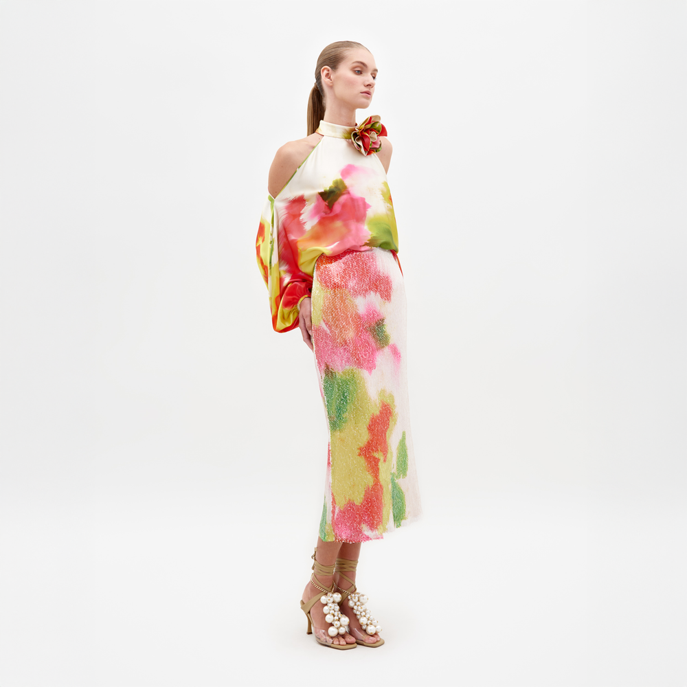 Una mujer está de pie sobre un fondo blanco vistiendo la Falda Sarla, una colorida falda midi con estampado floral hecha de textil italiano, combinada con un top halter a juego y sandalias adornadas con perlas color canela.