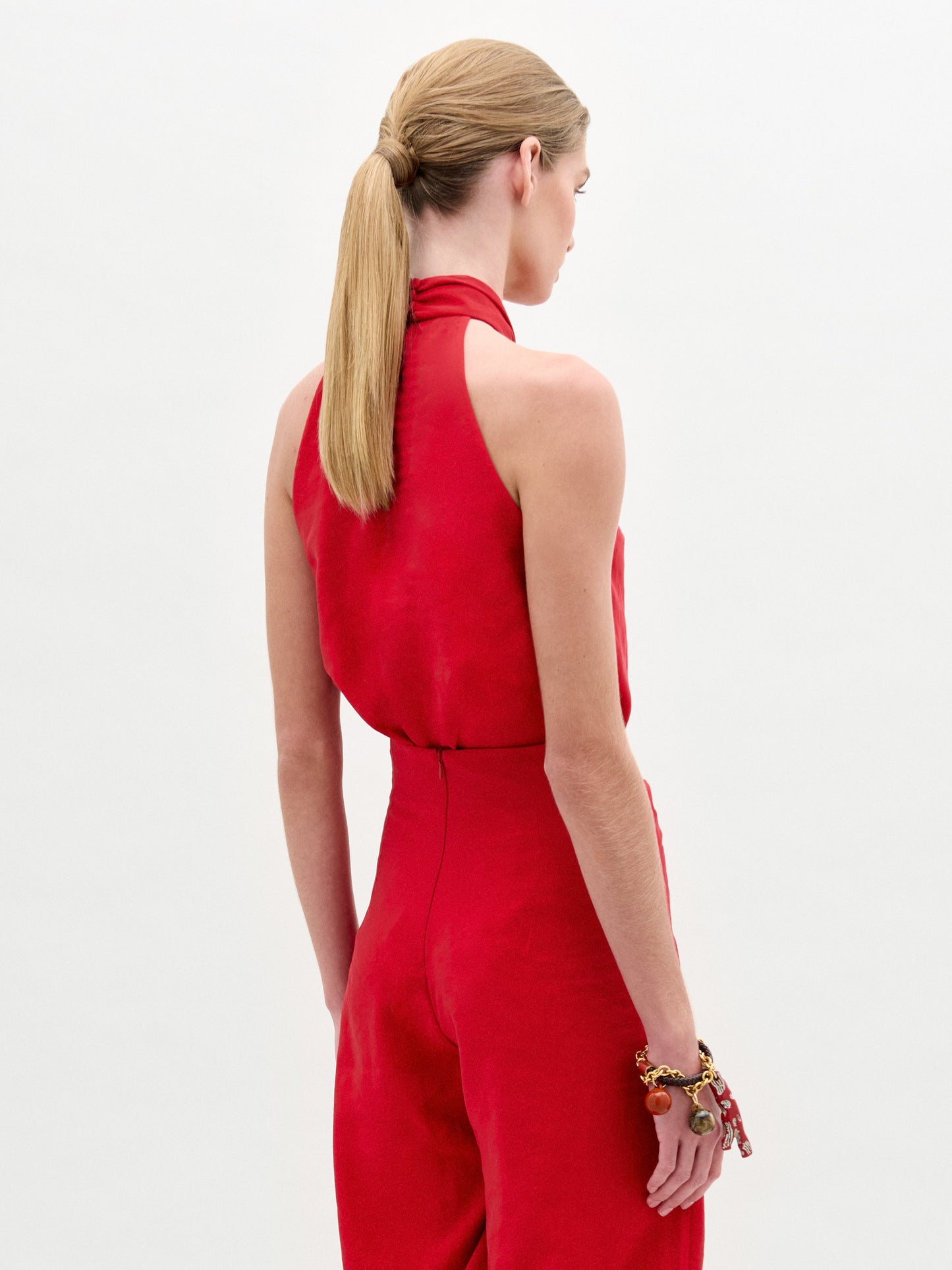Una persona con el pelo castaño claro y liso recogido en una coleta baja se ve de espaldas, llevando la blusa roja sin mangas y de cuello alto Shela Blouse hecho en Colombia y una pulsera gruesa, de pie sobre un fondo blanco liso.