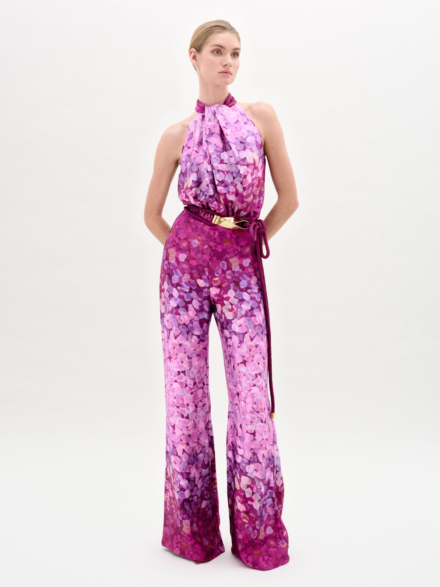 Una persona de pie sobre fondo blanco lleva el mono Siv Jumpsuit con estampado abstracto rosa y morado, pantalón de pata ancha y cinturón a juego con hebilla dorada, fabricado en Colombia.