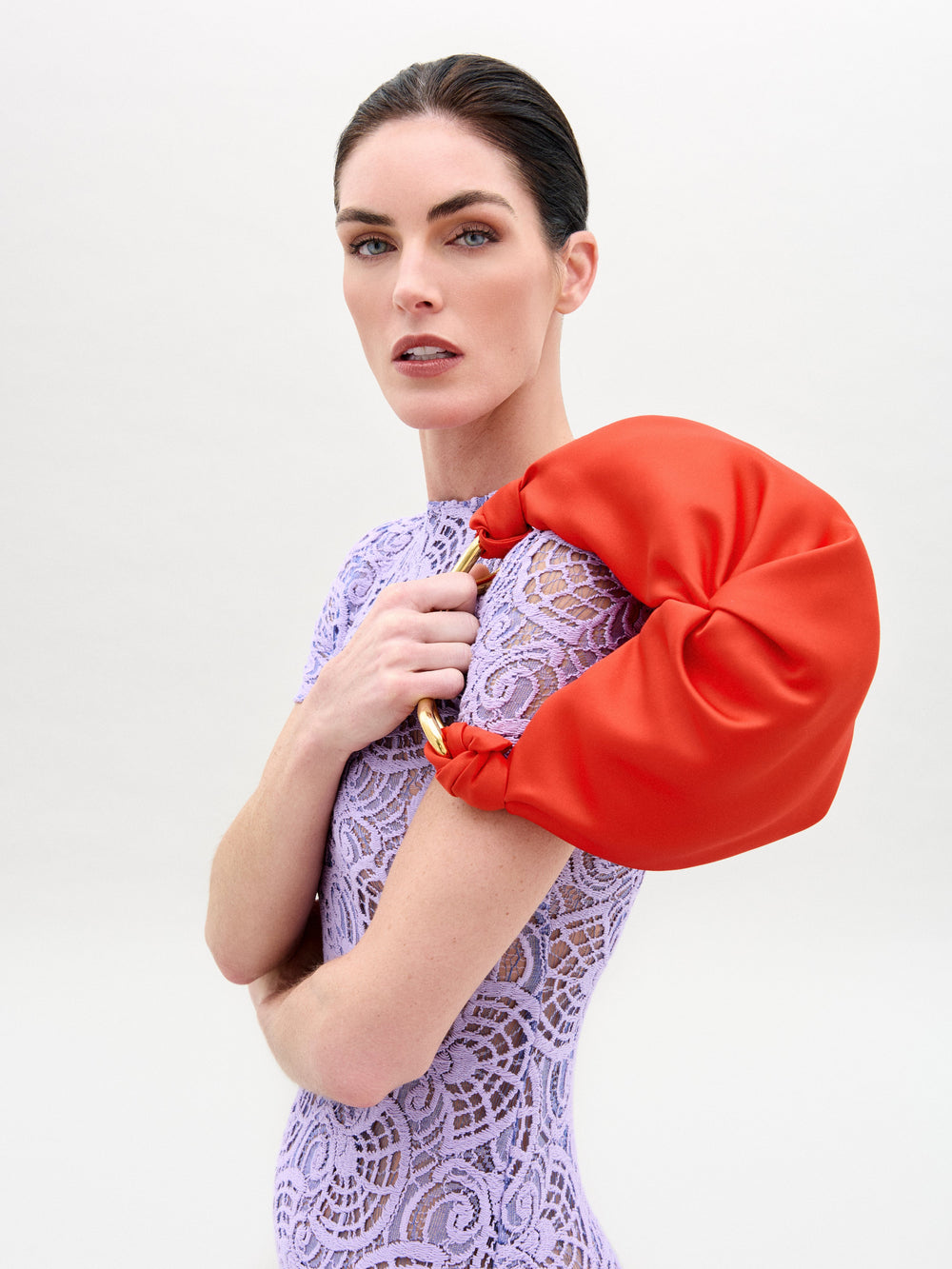 Una persona de cabello oscuro se encuentra de pie frente a un fondo liso, con un vestido de encaje color lavanda y sosteniendo el Bolso Sofia Small sobre su hombro. Fabricado en Colombia, este pequeño bolso rojo le agrega estilo a su expresión neutral mientras posa con la cabeza ligeramente inclinada.