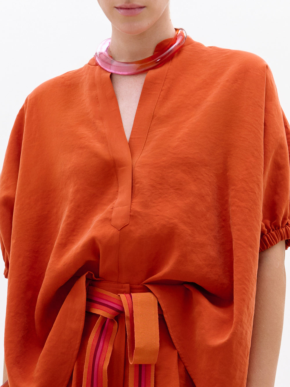 Una persona lleva la blusa Susane: una túnica naranja con cuello en V y mangas cortas fruncidas, un cinturón de rayas multicolor y una gruesa gargantilla rosa translúcida. Fabricada en Colombia. El fondo es liso y de color claro.
