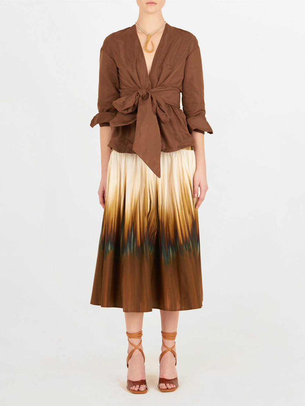 Una persona de pie sobre un fondo blanco lleva el Blusa Saanvi Brown, una falda midi ombre marrón y crema, tacones de tiras color canela y un collar con colgante. No se ve la cara.