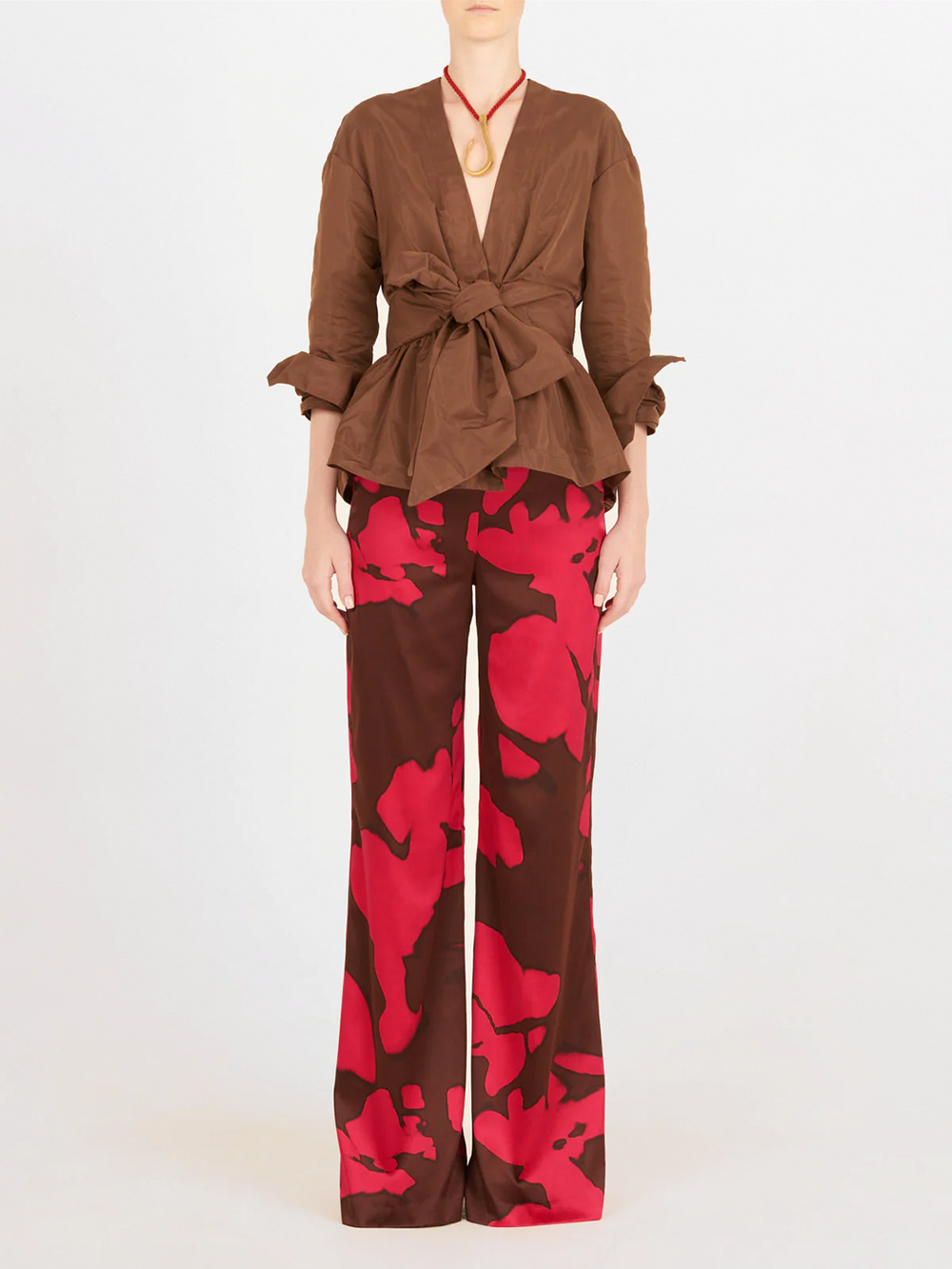 Una persona aparece sobre un fondo liso con un top marrón con lazada delantera y manga larga, combinado con el pantalón Andie Fucshia Cacao, con un atrevido estampado abstracto en rojo y marrón.