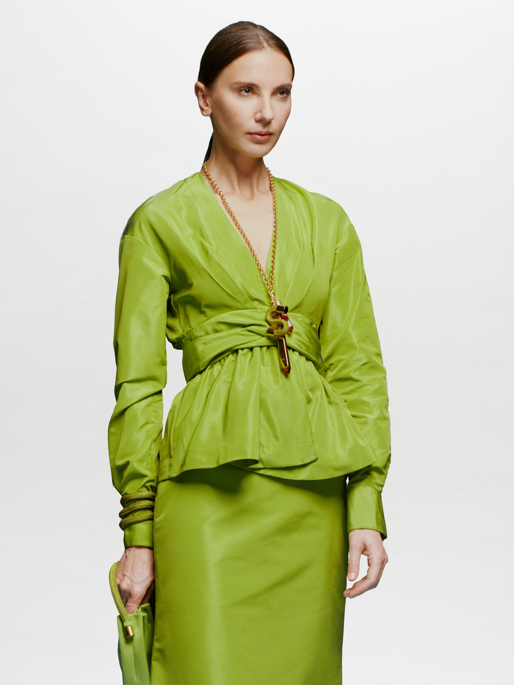 Una persona que viste una blusa de manga larga Blusa Saanvi de color verde brillante y una falda a juego está parada sobre un fondo blanco, sosteniendo un bolso de mano verde y luciendo un collar de cadena dorada.