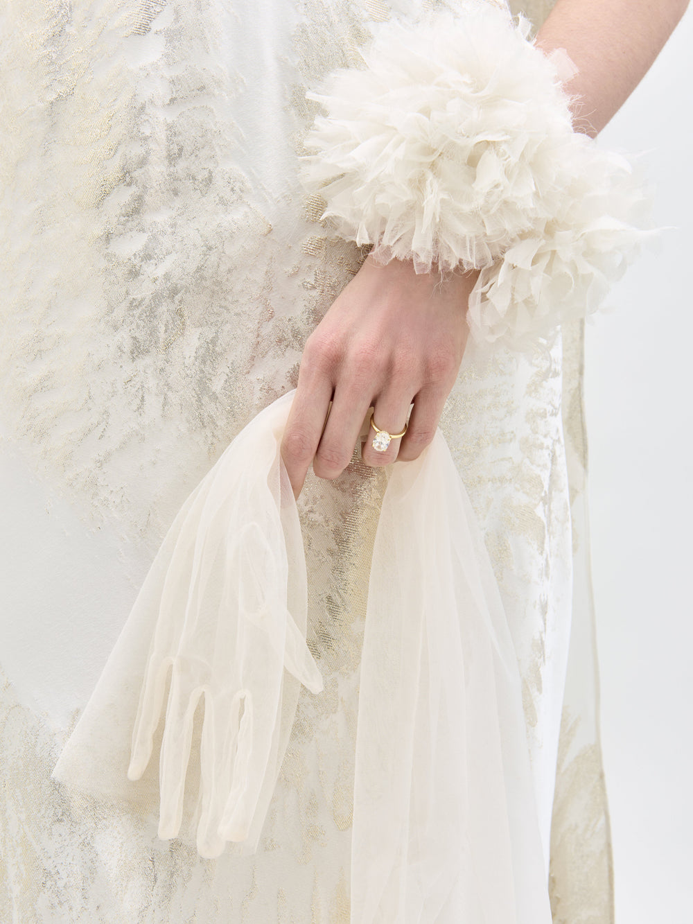 Una persona que viste el Vestido Sabina, una prenda blanca texturizada, sostiene guantes blancos transparentes en una mano. Su muñeca luce un puño blanco y esponjoso, y lleva un anillo de oro con una piedra blanca en un dedo.