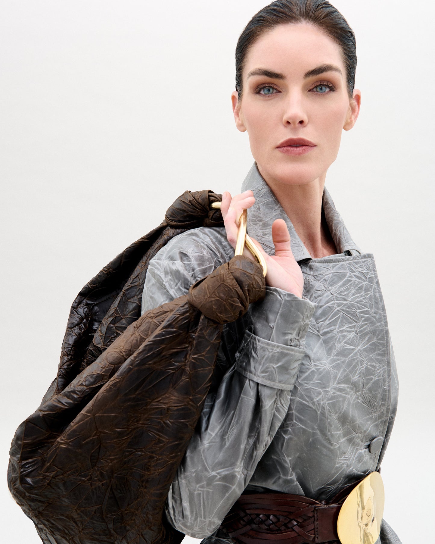 Una mujer de ojos claros y pelo oscuro recogido lleva un abrigo gris texturizado, un cinturón marrón ancho con hebilla dorada y lleva la Bolsa Sofía (Maxi) al hombro sobre un fondo liso.