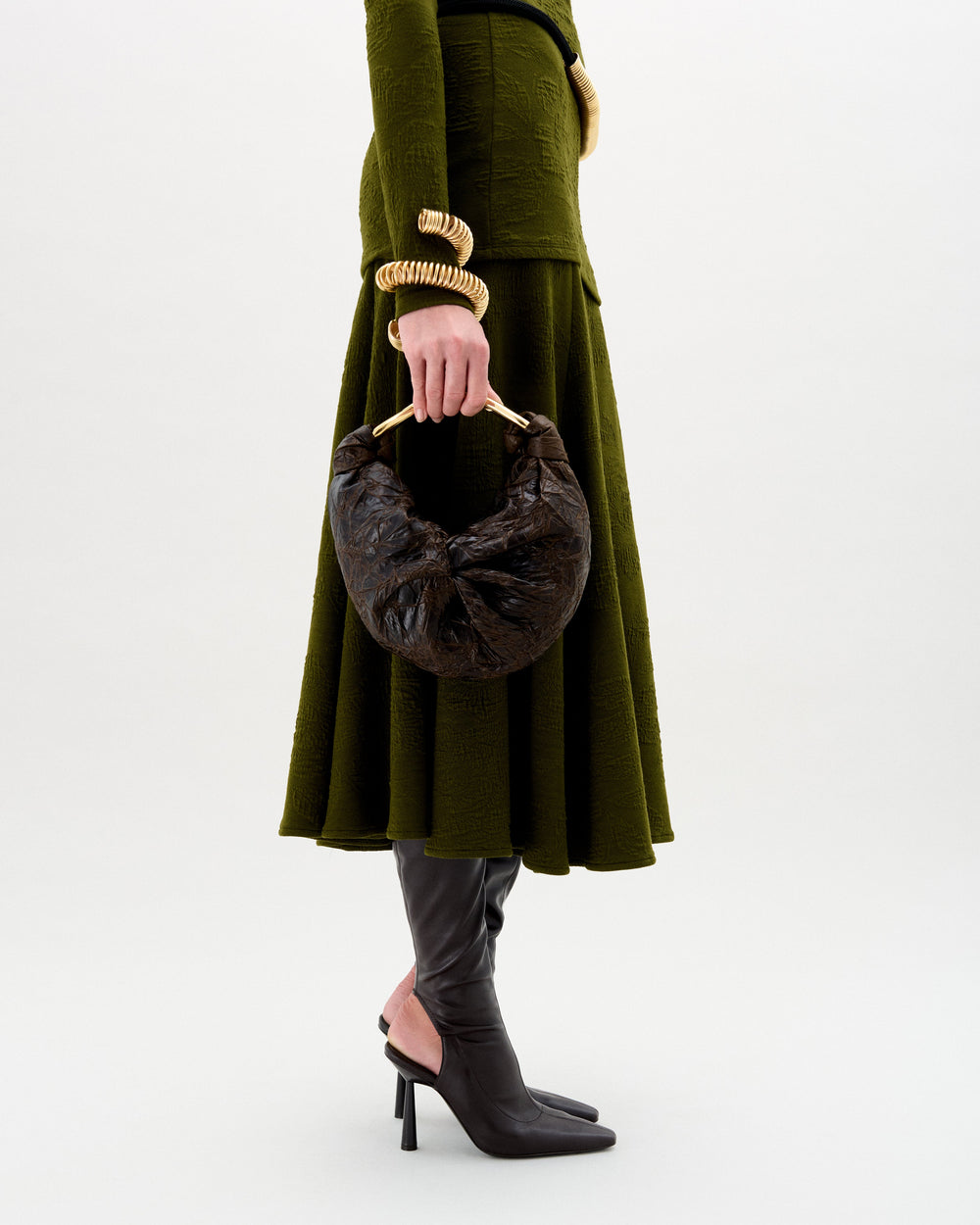 Una persona lleva un vestido verde oliva texturizado, botas negras de tacón alto, joyas doradas y el bolso Sofía (pequeño), un bolso de mano marrón oscuro con forma de media luna y asa redondeada, sobre un fondo liso.