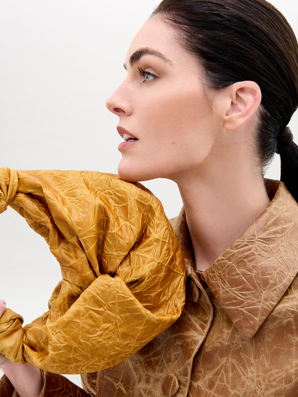 Una persona de perfil, con el cabello oscuro recogido, lleva un abrigo marrón texturizado. Sostiene el Bolso Sofia Small, un accesorio arrugado de tela italiana color mostaza, cerca del cuello sobre un fondo liso.