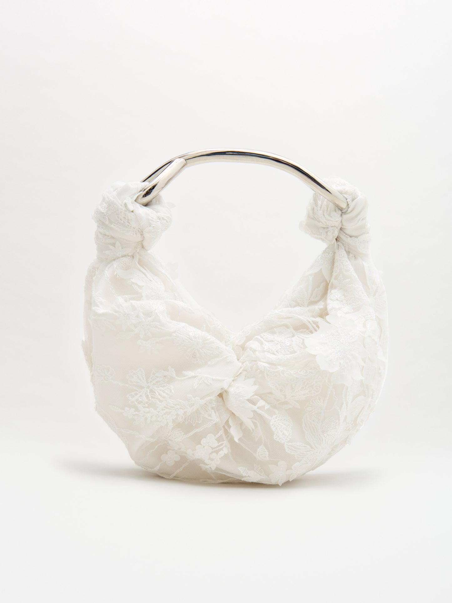 El Bolso Sofía es un bolso blanco con una capa de encaje floral y un llamativo nudo en la base de su asa curva de metal plateado. Se presenta sobre un fondo blanco liso.