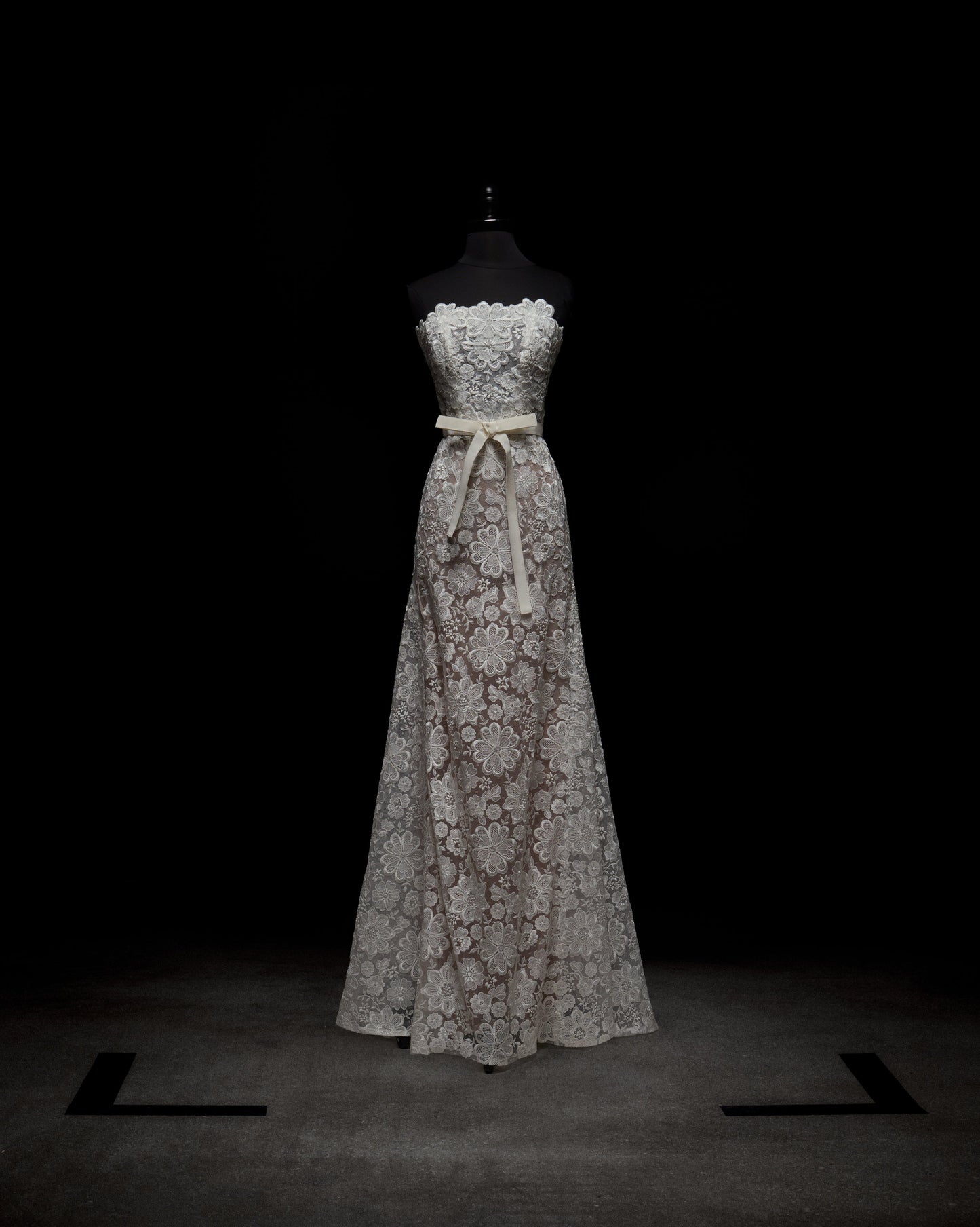 El Vestido Valencia es un vestido largo, sin tirantes, de encaje blanco con escote festoneado y una cinta ligera en la cintura, exhibido en un maniquí negro sobre un fondo oscuro.