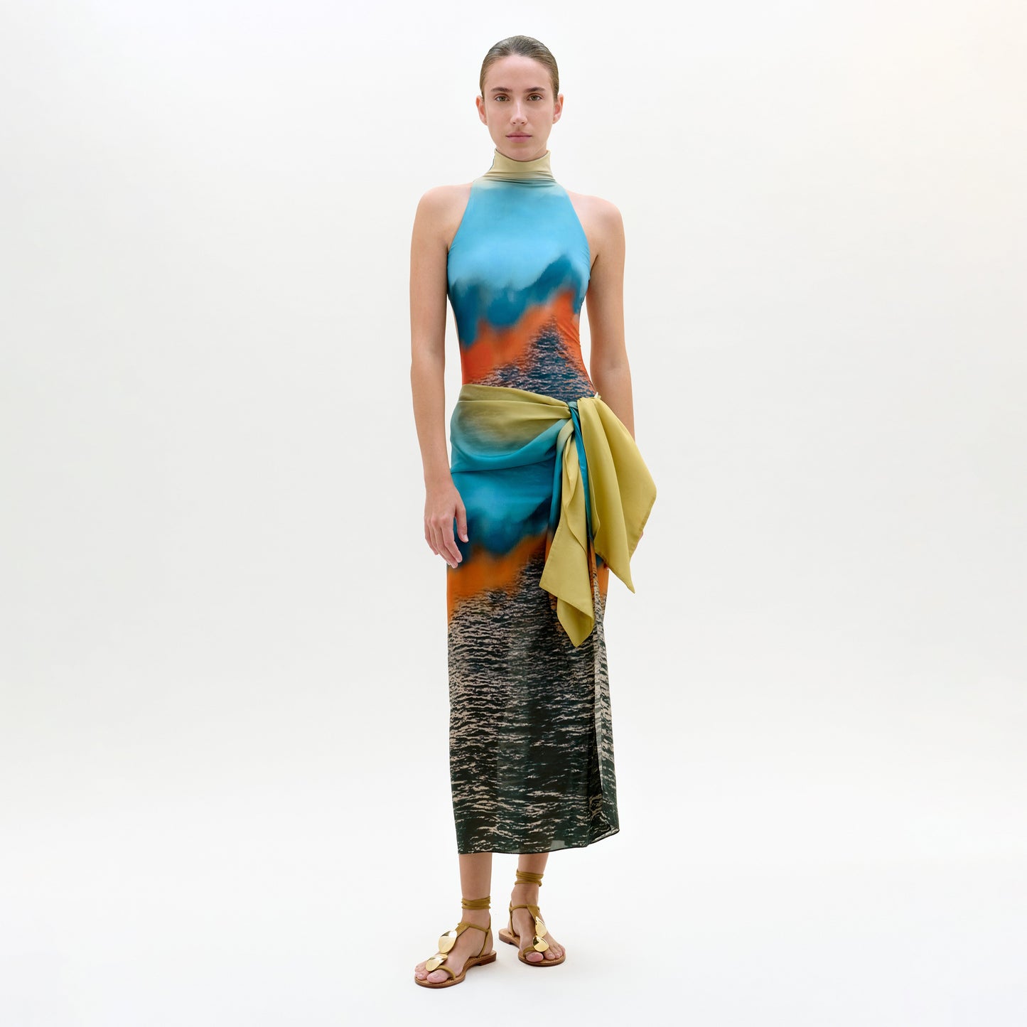 Una mujer de frente con el Viveka Pareo, vestido sin mangas de cuello alto con estampado de agua y puesta de sol, fajín verde-amarillo en la cintura, falda de largo midi y sandalias planas doradas sobre un fondo claro liso.