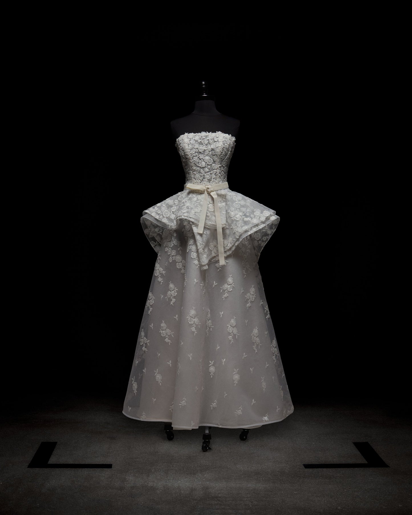 El Vestido Viviana es un vestido blanco sin tirantes con un corpiño de encaje ajustado, una sobrefalda estilo peplum, un cinturón de cinta en la cintura y una falda larga de encaje semitransparente, que se muestra en un maniquí negro sobre un fondo oscuro.