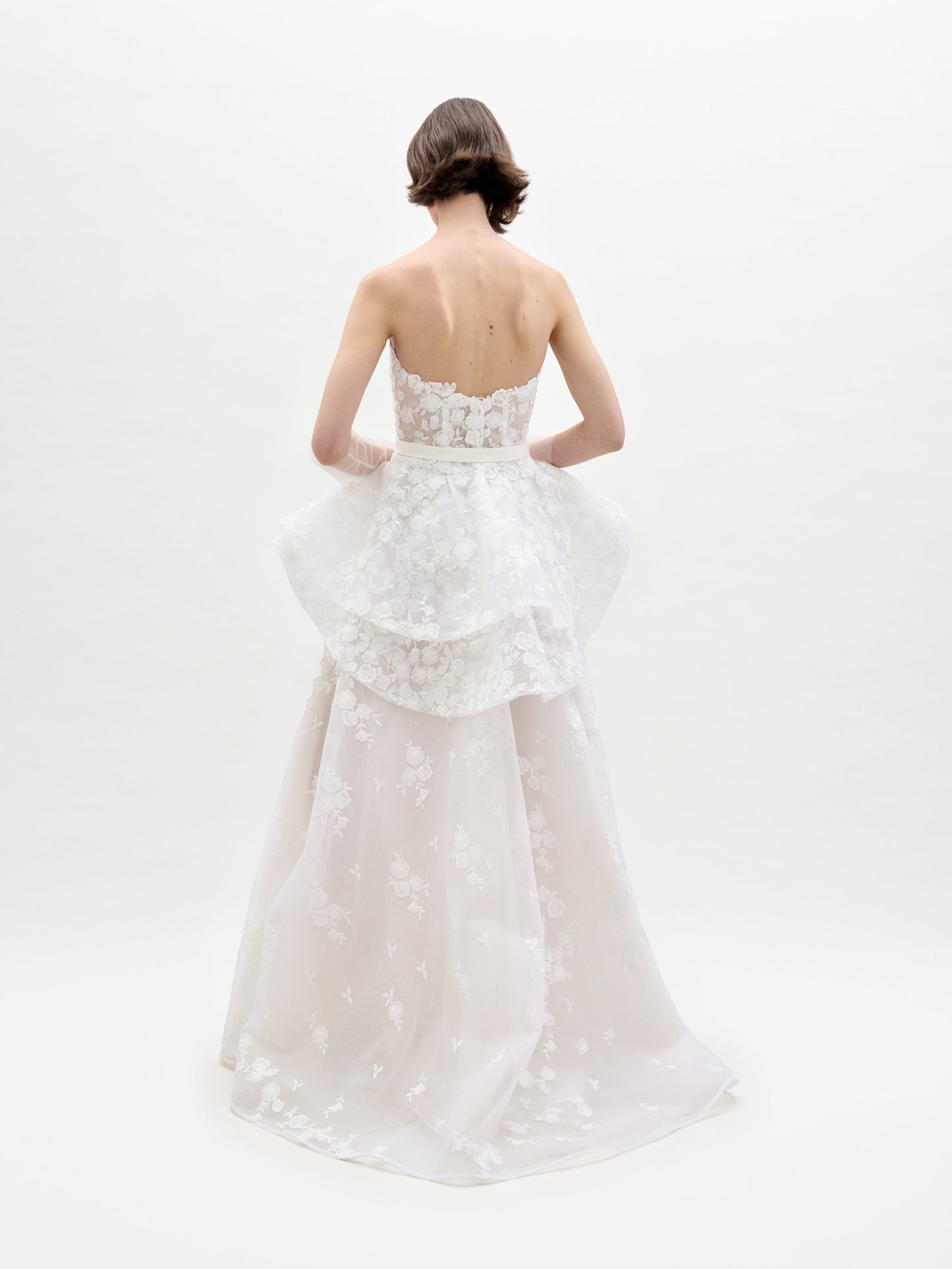 Una persona con cabello castaño corto mira hacia otro lado, modelando el Vestido Viviana, un vestido blanco sin tirantes con bordados florales y cintura peplum, sobre un fondo claro y liso.