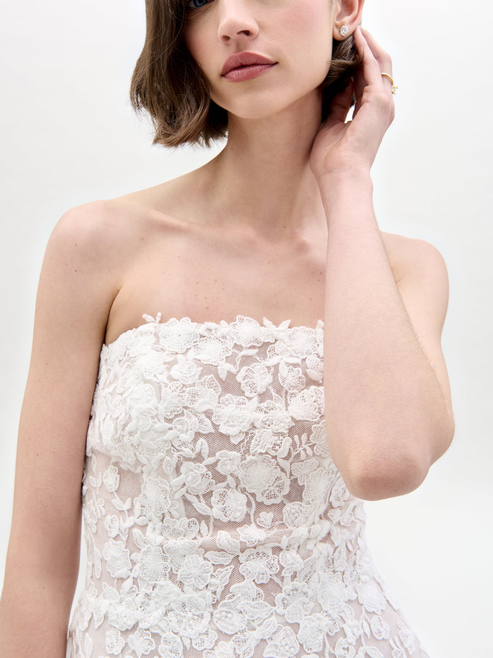 Una mujer de cabello castaño corto luce el Vestido Viviana, un vestido de encaje blanco sin tirantes. Se toca la oreja con una mano, de pie sobre un fondo claro y liso, con el rostro parcialmente fuera del encuadre.