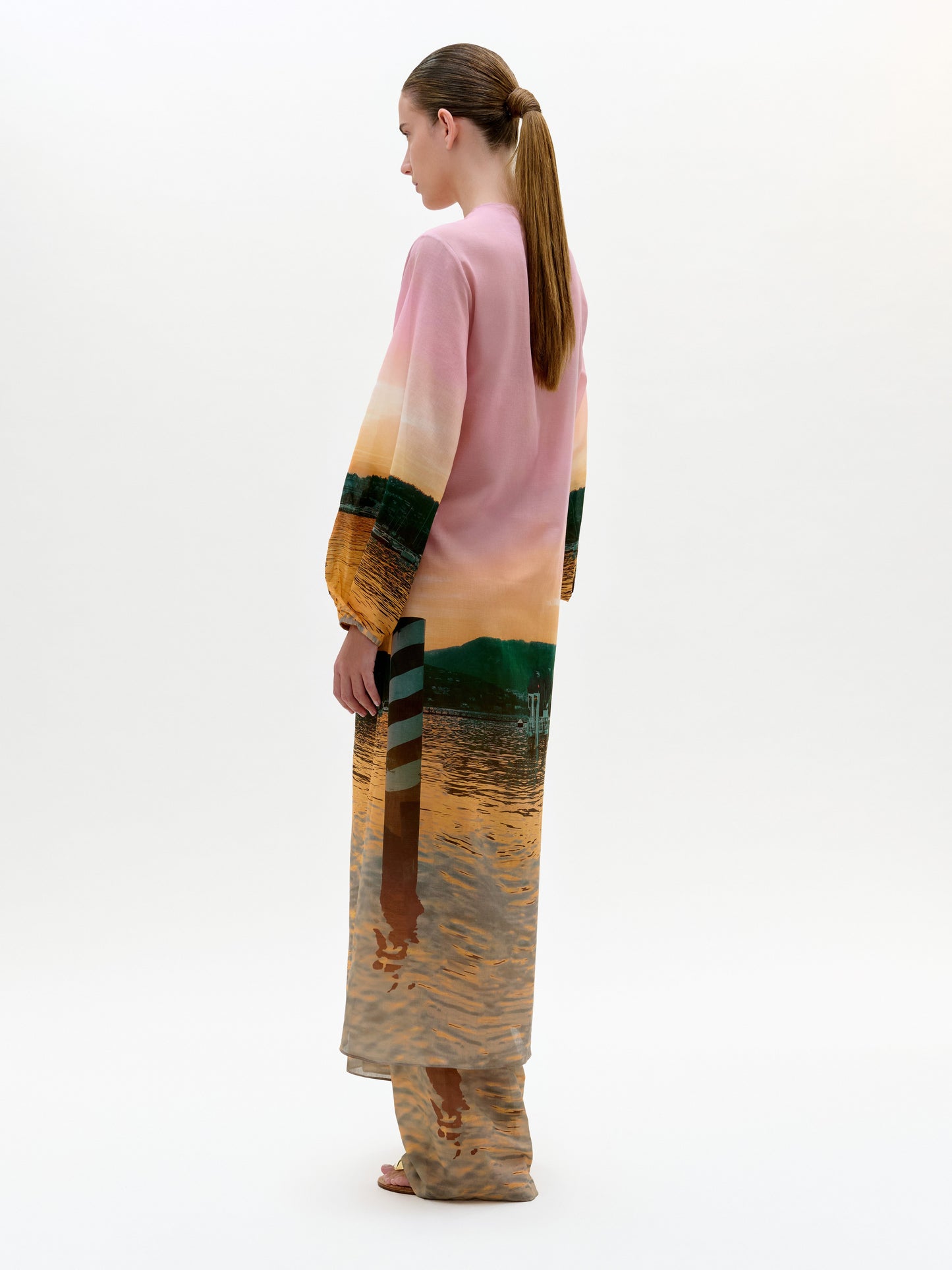 Una mujer de perfil luce la bata Yanil, un vestido manga larga 100% algodón con estampado de agua y árboles lejanos al atardecer. Cálidos tonos rosas, naranjas y verdes resaltan su elegante escote en V profundo. Lleva el pelo recogido en una coleta baja.