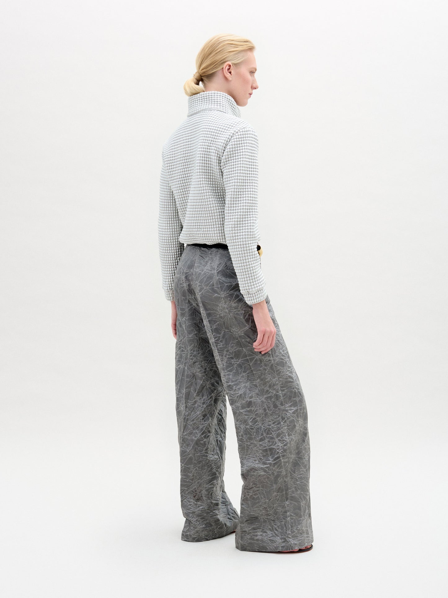 Una persona con el pelo rubio recogido, vista de espaldas, luce el pantalón Emine Pant, un pantalón gris de tiro alto y pierna ancha fabricado en Colombia, combinado con un top blanco texturizado, todo ello sobre un fondo blanco liso.