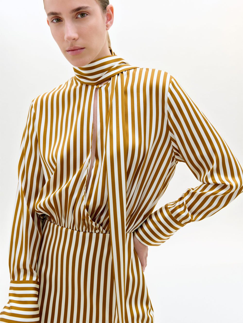 Una persona lleva el vestido Imelda, de rayas verticales doradas y blancas en lujosa seda, con mangas largas, escote alto y detalle de recorte delantero. El fondo es liso y claro. Fabricado en Portugal.