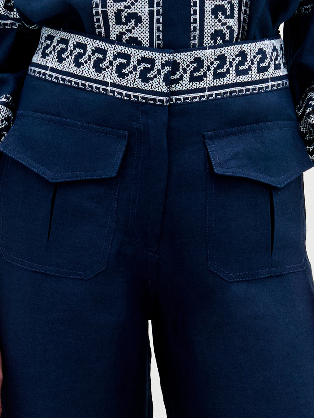 Primer plano de una persona que lleva el pantalón Zina, de lino azul oscuro, con grandes bolsillos delanteros con solapa y perneras anchas. También se ve un cinturón estampado, una cintura alta y parte de un top bordado a juego.