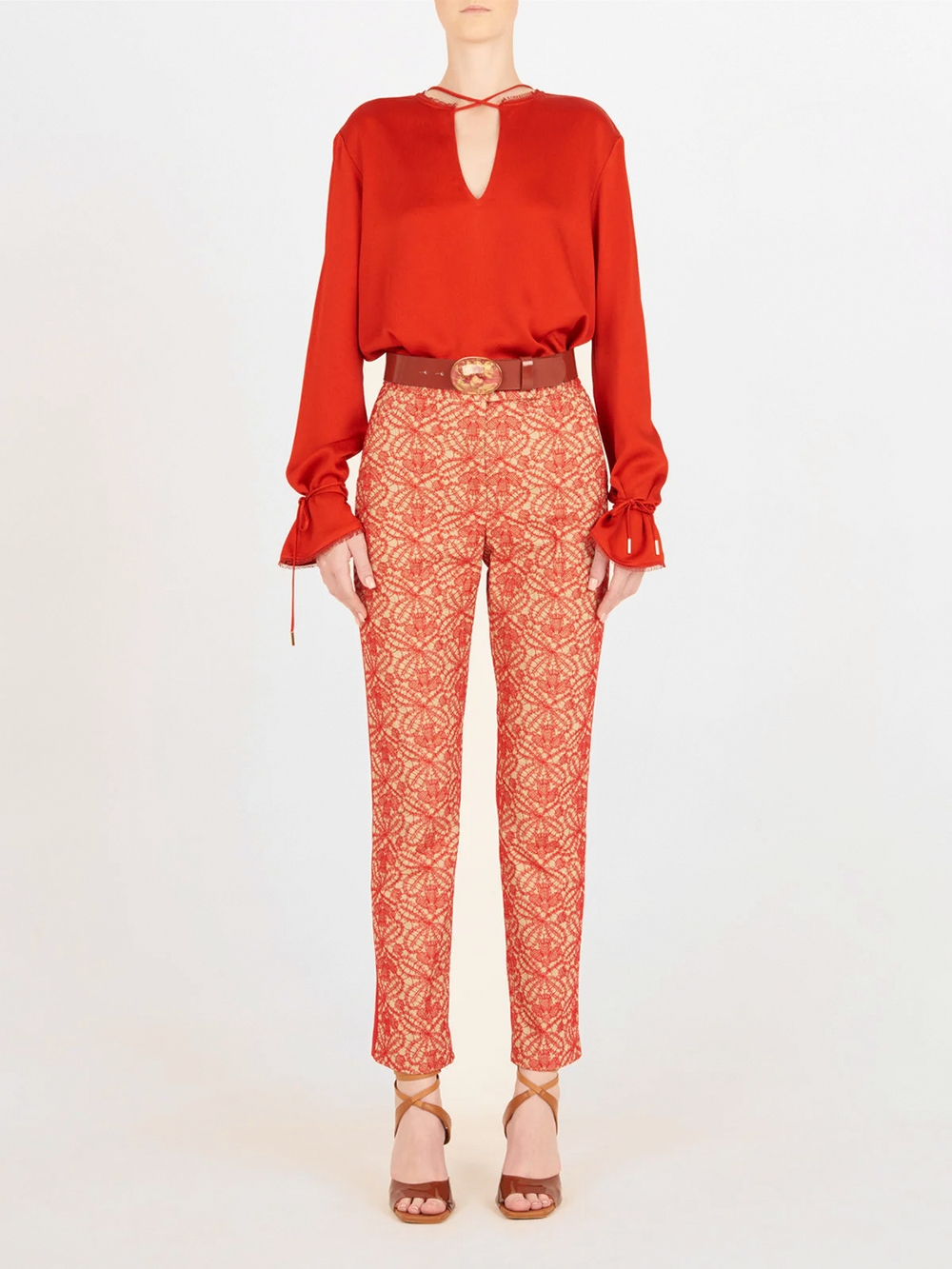 Una persona de pie sobre un fondo liso lleva un Blusa Zafra Rouge de manga larga, pantalones rojos y beige estampados, sandalias marrones de tacón alto y un cinturón marrón con hebilla redonda.
