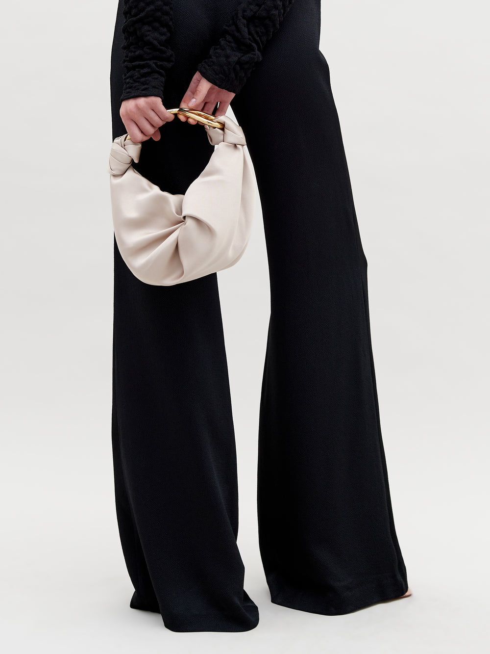 Una persona que viste una blusa negra texturizada de manga larga y pantalones anchos sostiene el Bolso Sofia Small, con herraje dorado y asa curva. El diseño tiene un fondo claro y sencillo.