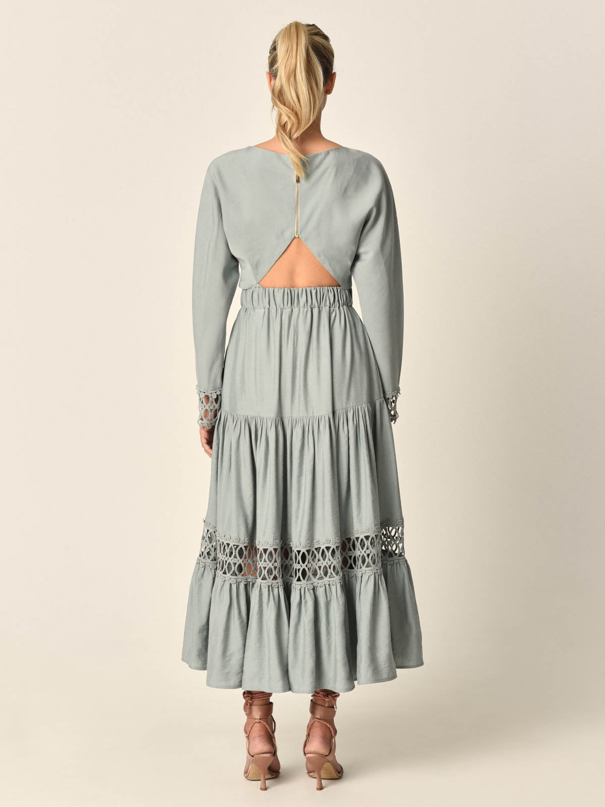 Una persona rubia con cola de caballo lleva el Top Celeste Sage, un top verde salvia claro de manga larga con aberturas en la espalda y paneles de encaje sobre el bajo. Lo combina con tacones rosas con cordones y se coloca de espaldas.