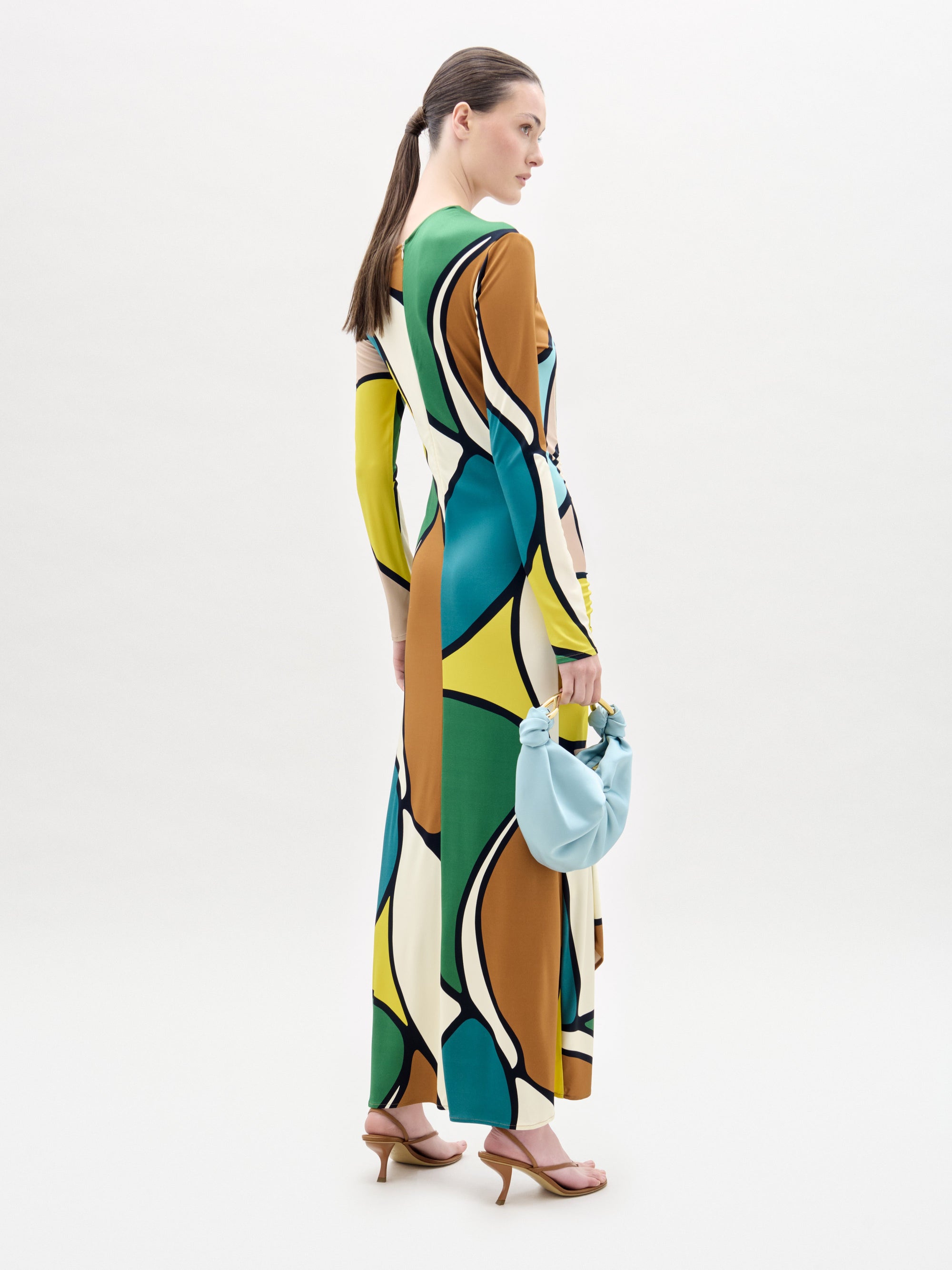 Una mujer con el pelo largo y castaño recogido en una coleta baja lleva el vestido Ilania, con formas verdes, amarillas, marrones, blancas y azules. Lleva un bolso azul claro y sandalias de tacón color canela sobre fondo blanco.