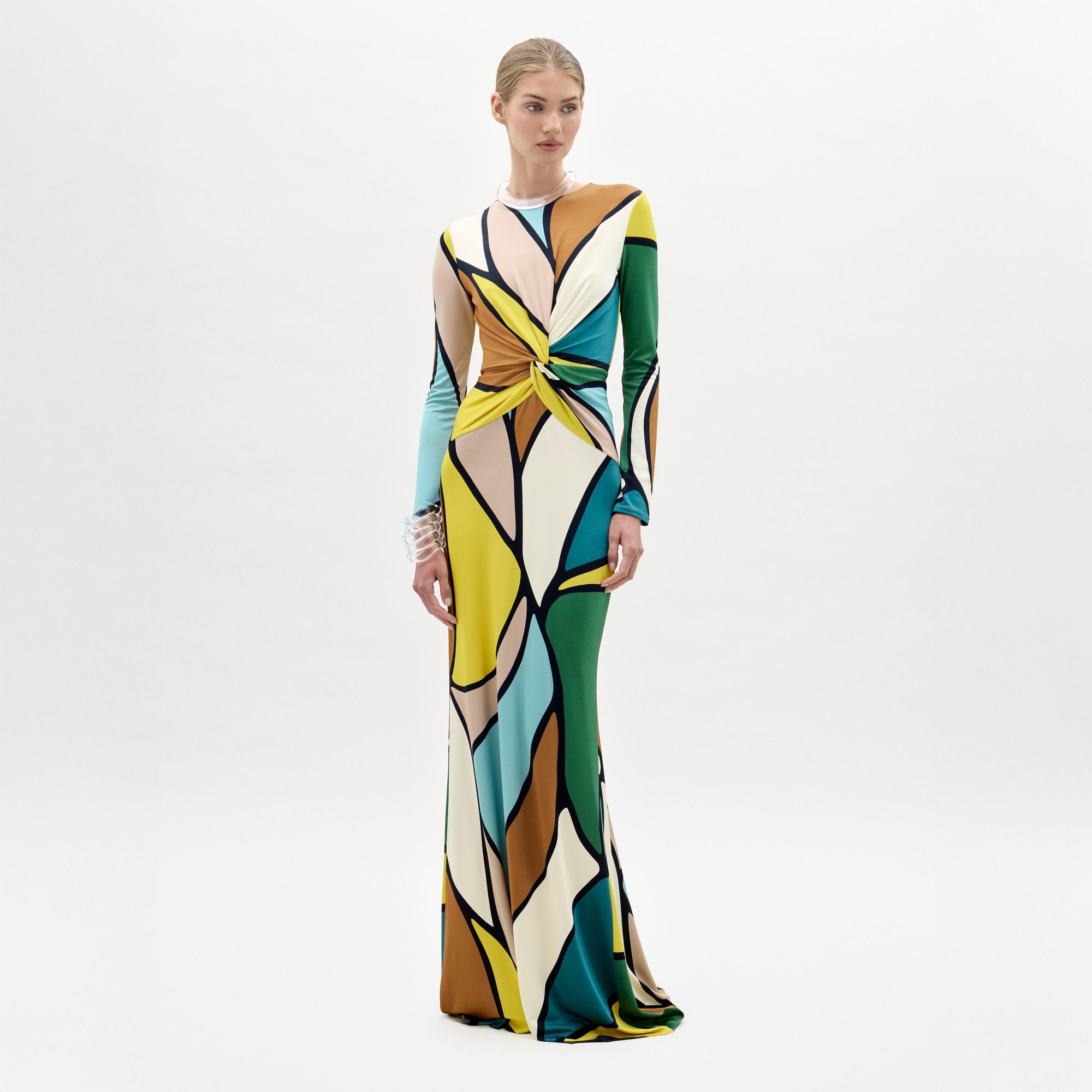 Una mujer, sobre un fondo blanco liso, lleva el vestido Lucian, un maxivestido de tela italiana con un colorido estampado abstracto, manga larga y un detalle enroscado en la cintura. Mira hacia delante con los brazos relajados a los lados.