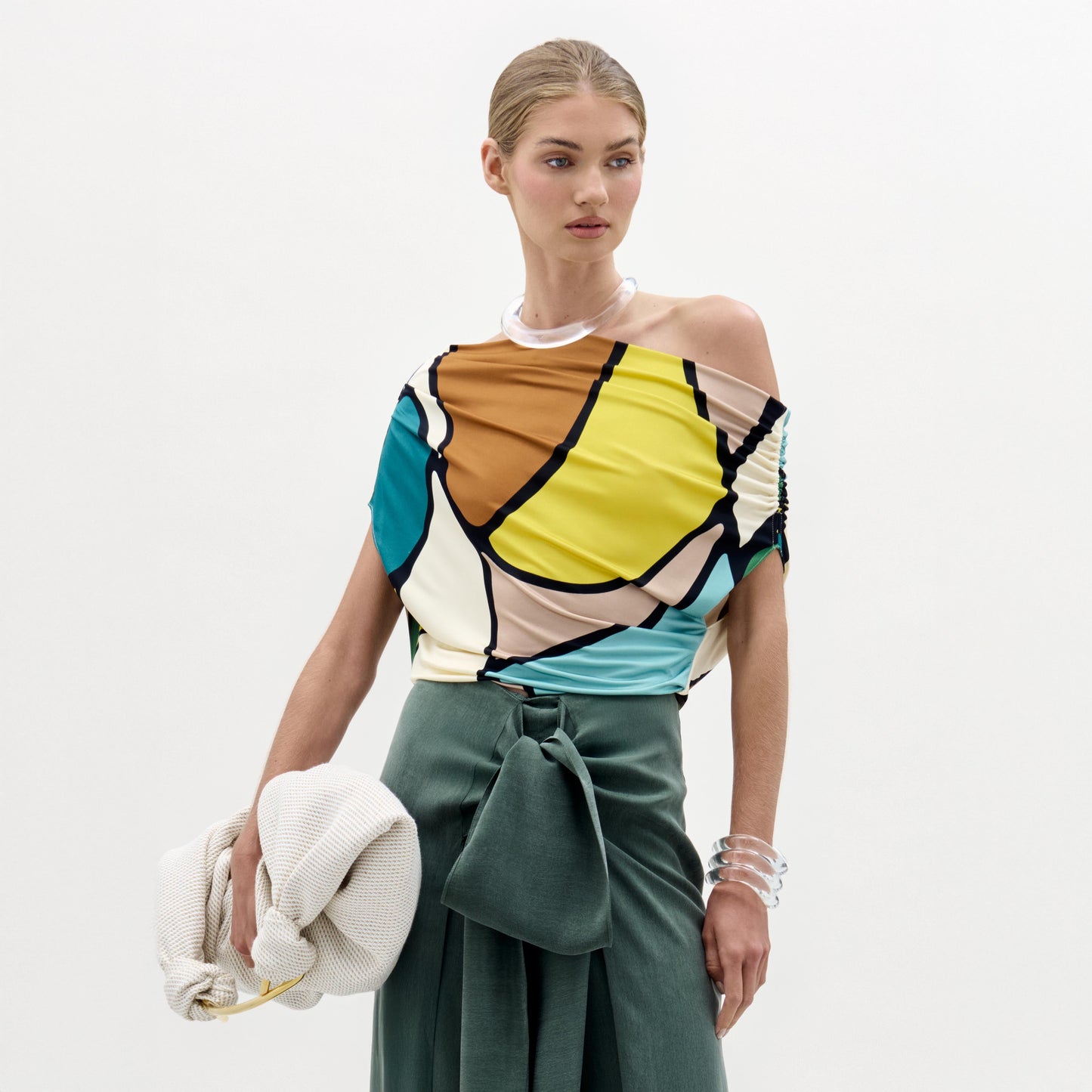Una mujer, sobre fondo blanco, lleva la colorida blusa Indiana -un top asimétrico de tejido italiano- con pantalones verdes de cintura alta y un gran lazo, sostiene un bolso blanco con textura y luce brazaletes translúcidos.