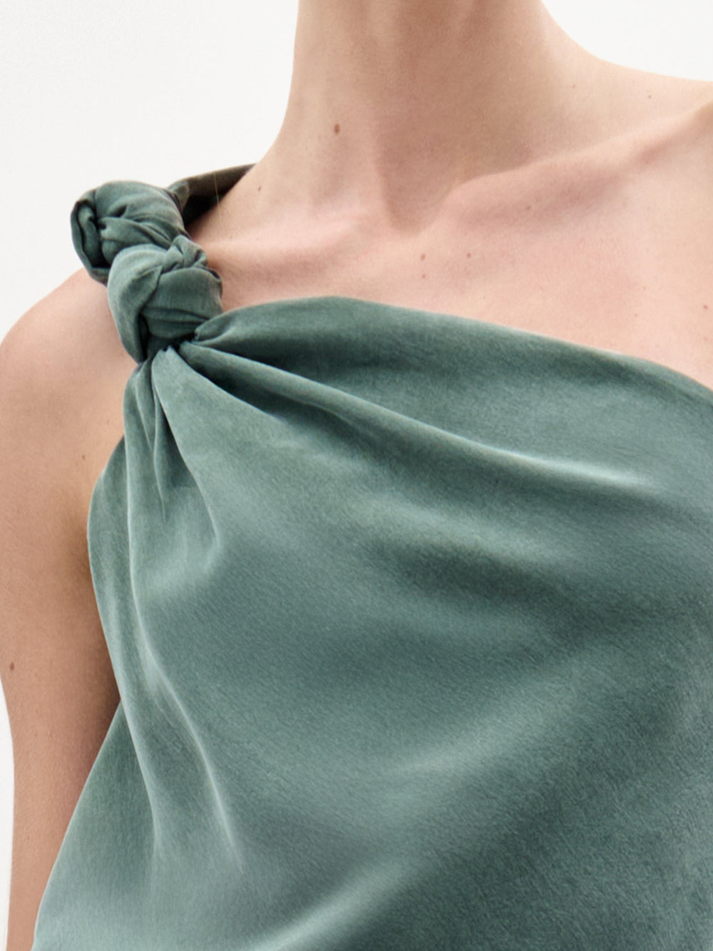 Primer plano de una persona que lleva la blusa Yanet, un top de seda verde salvia sin mangas, con escote asimétrico y un nudo en un hombro, sobre un fondo blanco minimalista.