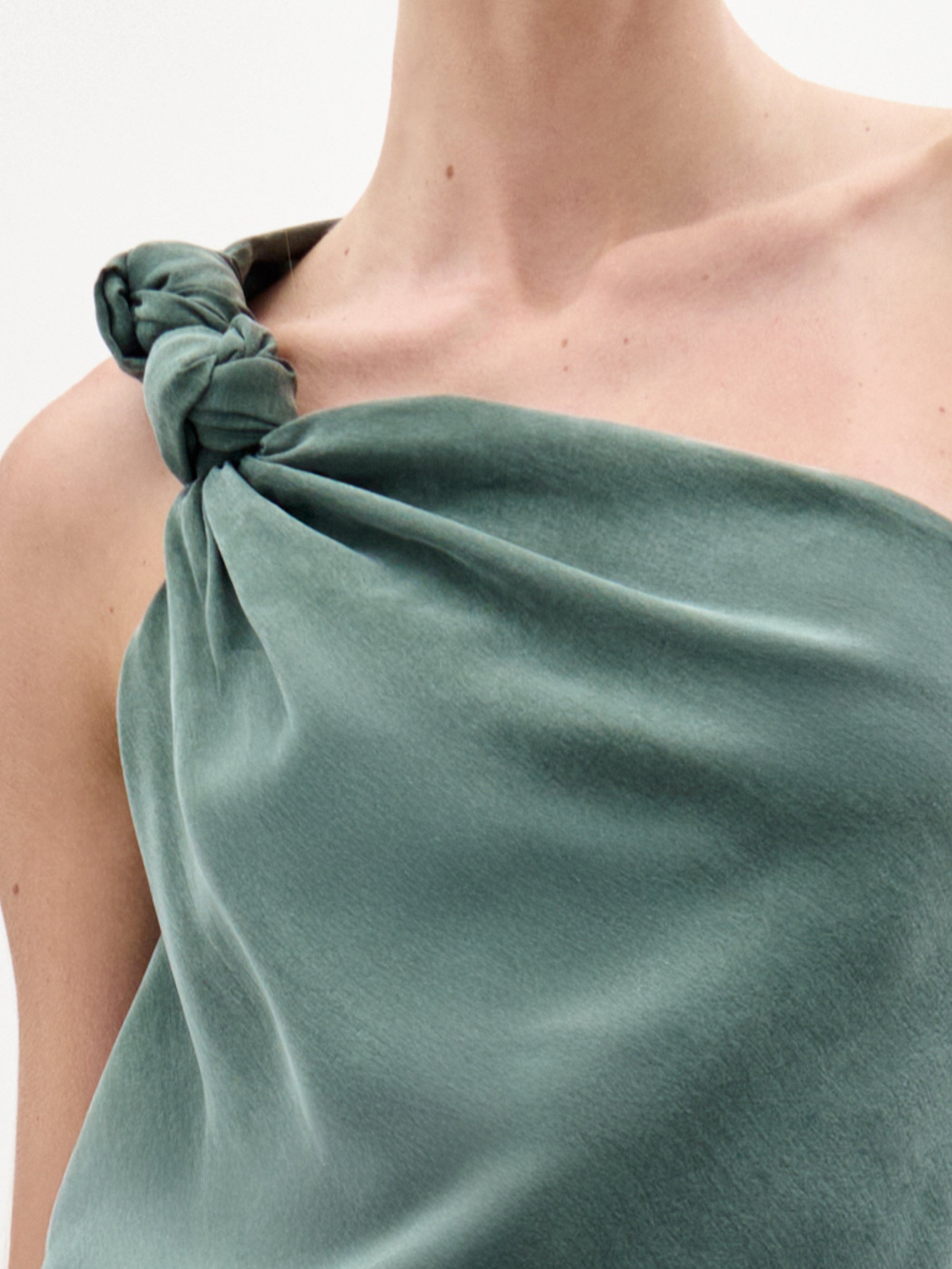 Primer plano de una persona que lleva la blusa Yanet, un top de seda verde salvia sin mangas, con escote asimétrico y un nudo en un hombro, sobre un fondo blanco minimalista.