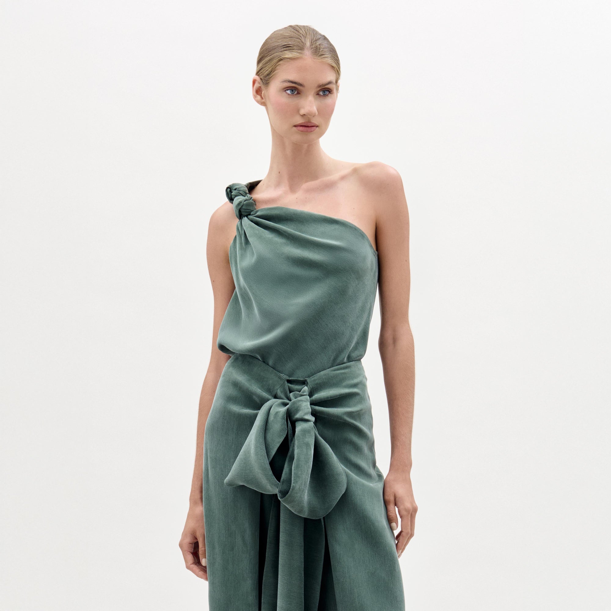 Una mujer de piel clara y pelo rubio recogido modela la blusa Yanet -seda verde salvia, escote asimétrico y un gran lazo en la cintura- sobre un fondo liso. Esta blusa está hecha en Colombia.