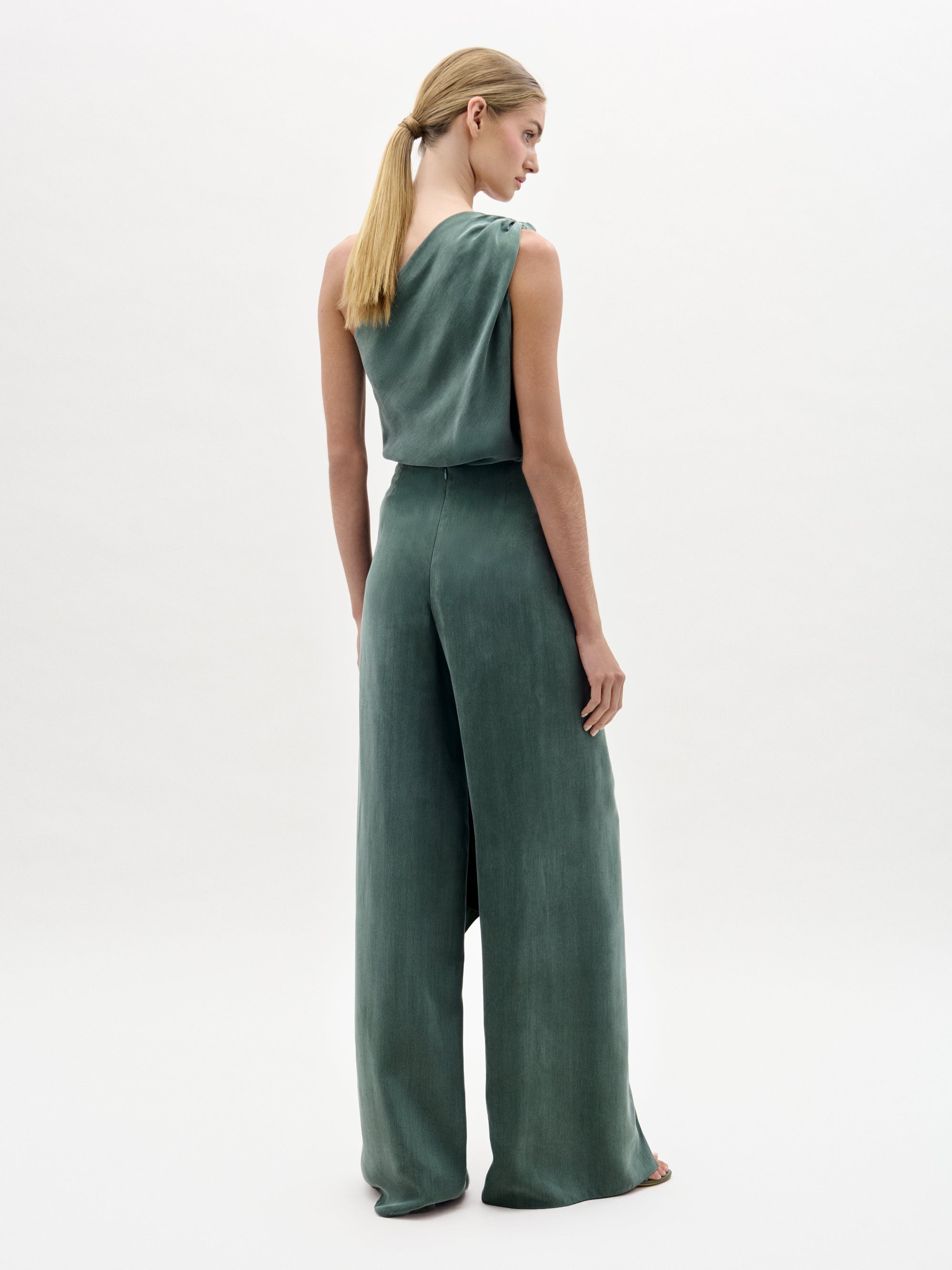 Una mujer con el pelo rubio recogido en una coleta baja está de espaldas, vestida con el Arenzano Pant -un mono verde salvia sin mangas confeccionado en tela italiana con pantalón largo y tiro alto- sobre un fondo liso y claro.