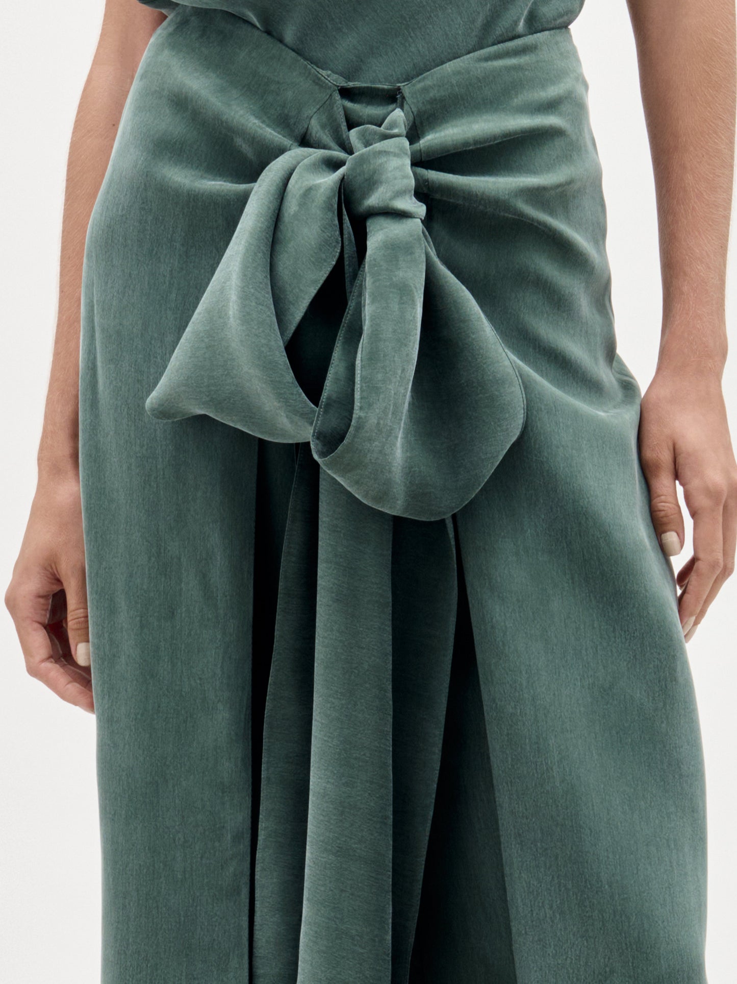 Una persona aparece de pie sobre un fondo liso, mostrando únicamente la parte inferior del torso y las manos, mientras lleva puesto el Arenzano Pant, un modelo verde, texturizado, de talle alto y con un gran detalle de nudo delantero.