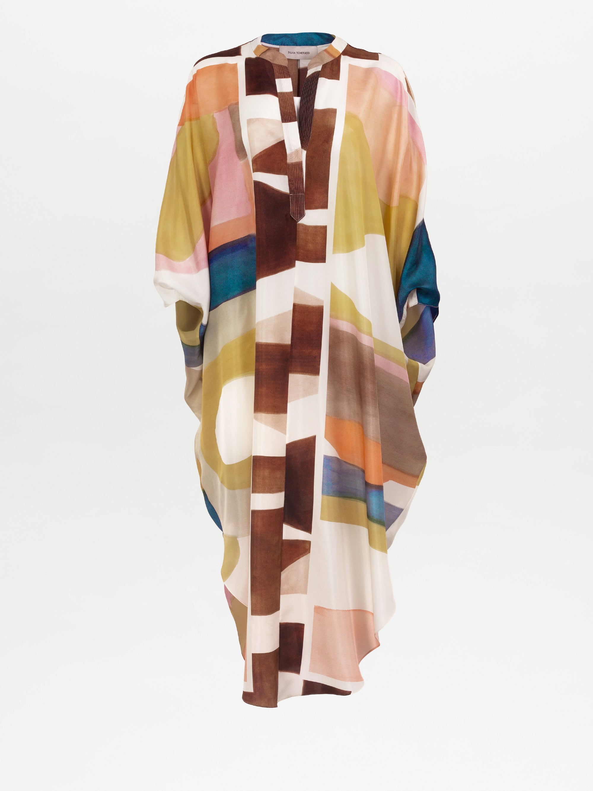 La túnica Ilse es un maxi caftán largo y holgado con mangas anchas, confeccionado en suave seda en Colombia. Presenta un estampado geométrico abstracto en marrón, rosa, amarillo, azul y blanco sobre un fondo claro con escote en V y cuello alto corto.